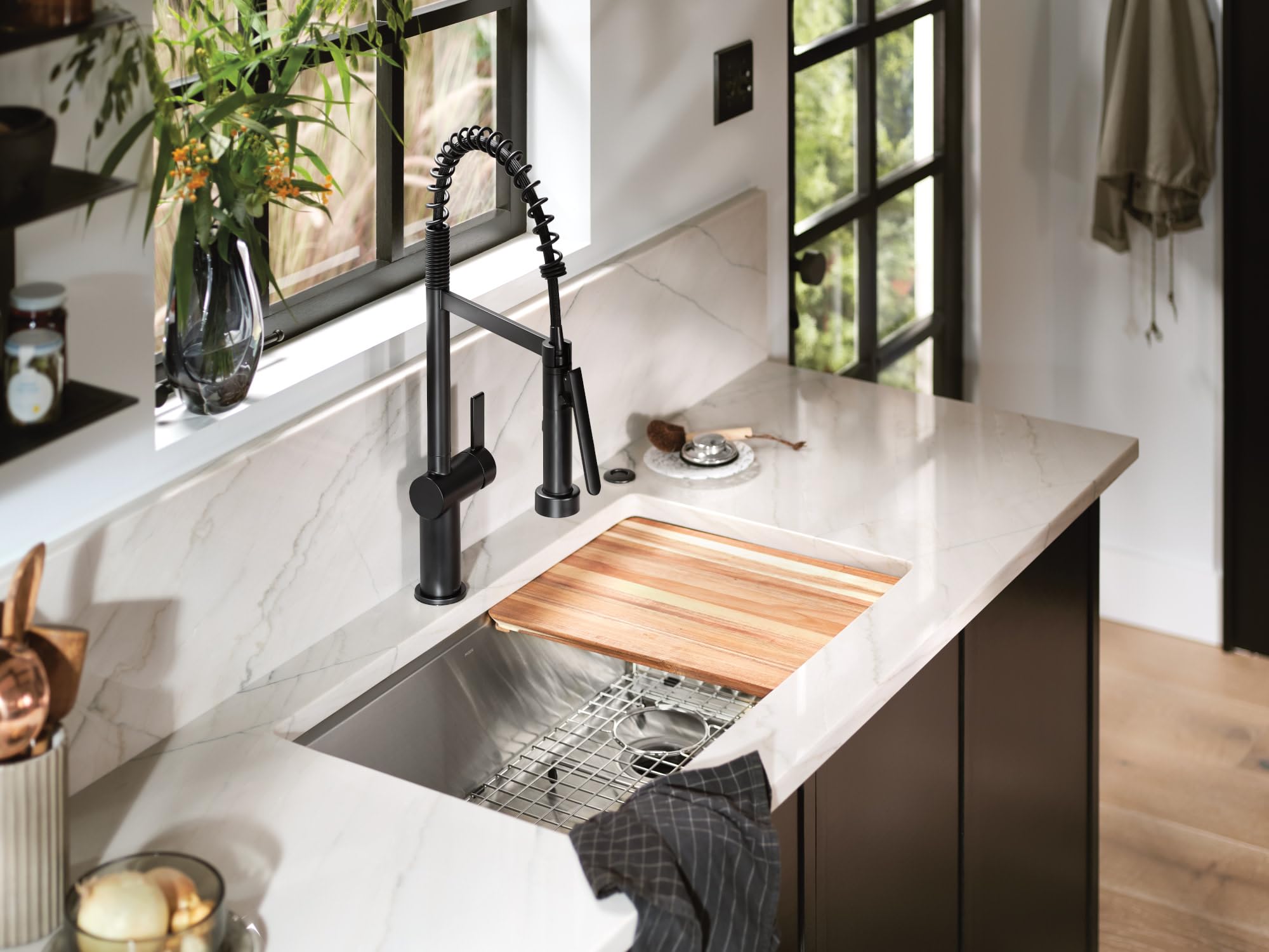 Matte black pre-rinse spring kitchen faucet with coiled hose installed above undermount sink with wooden cutting board.