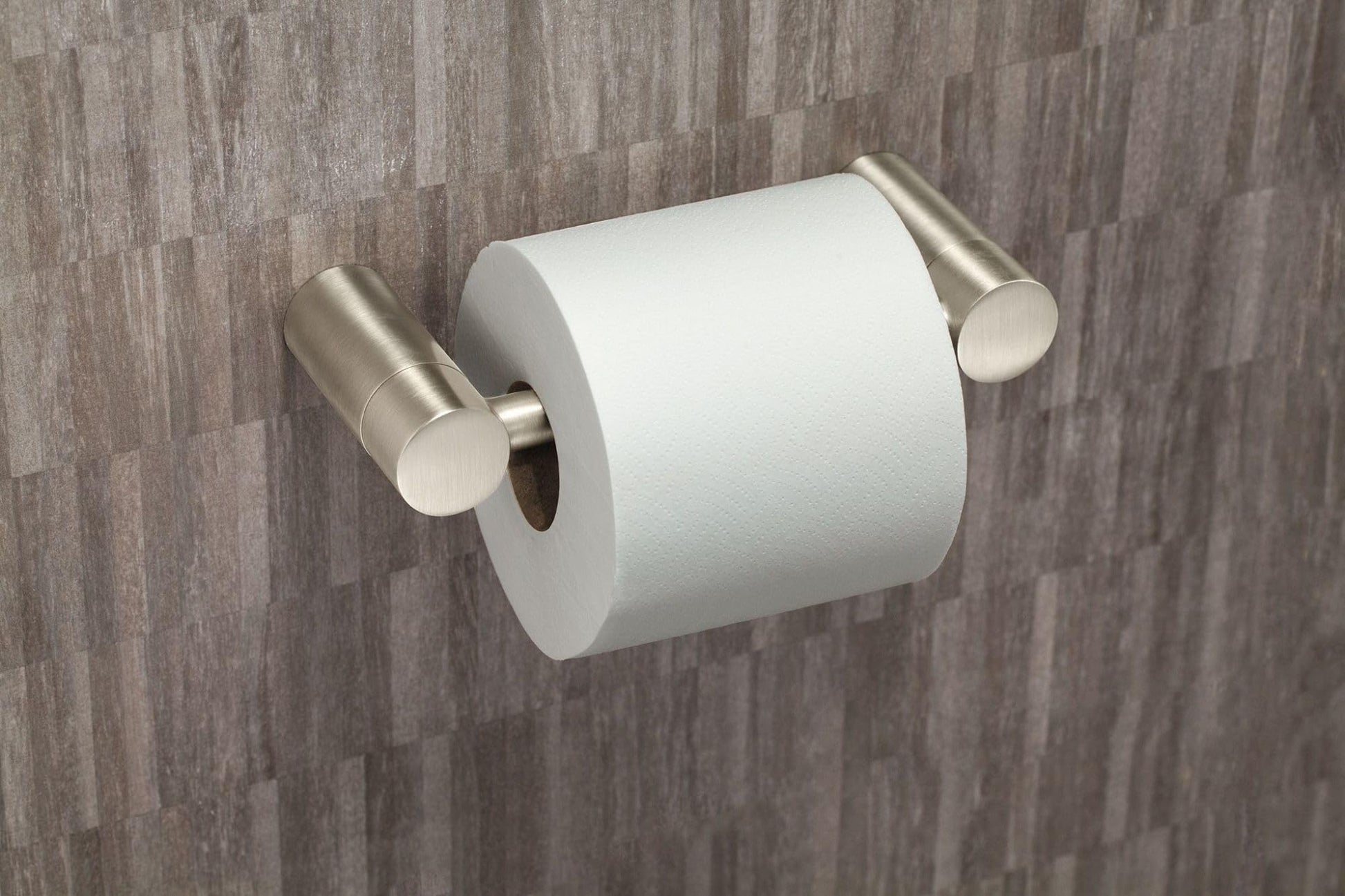 Brushed nickel toilet paper holder with cylindrical mounting hardware holding a white toilet paper roll against tiled wall.