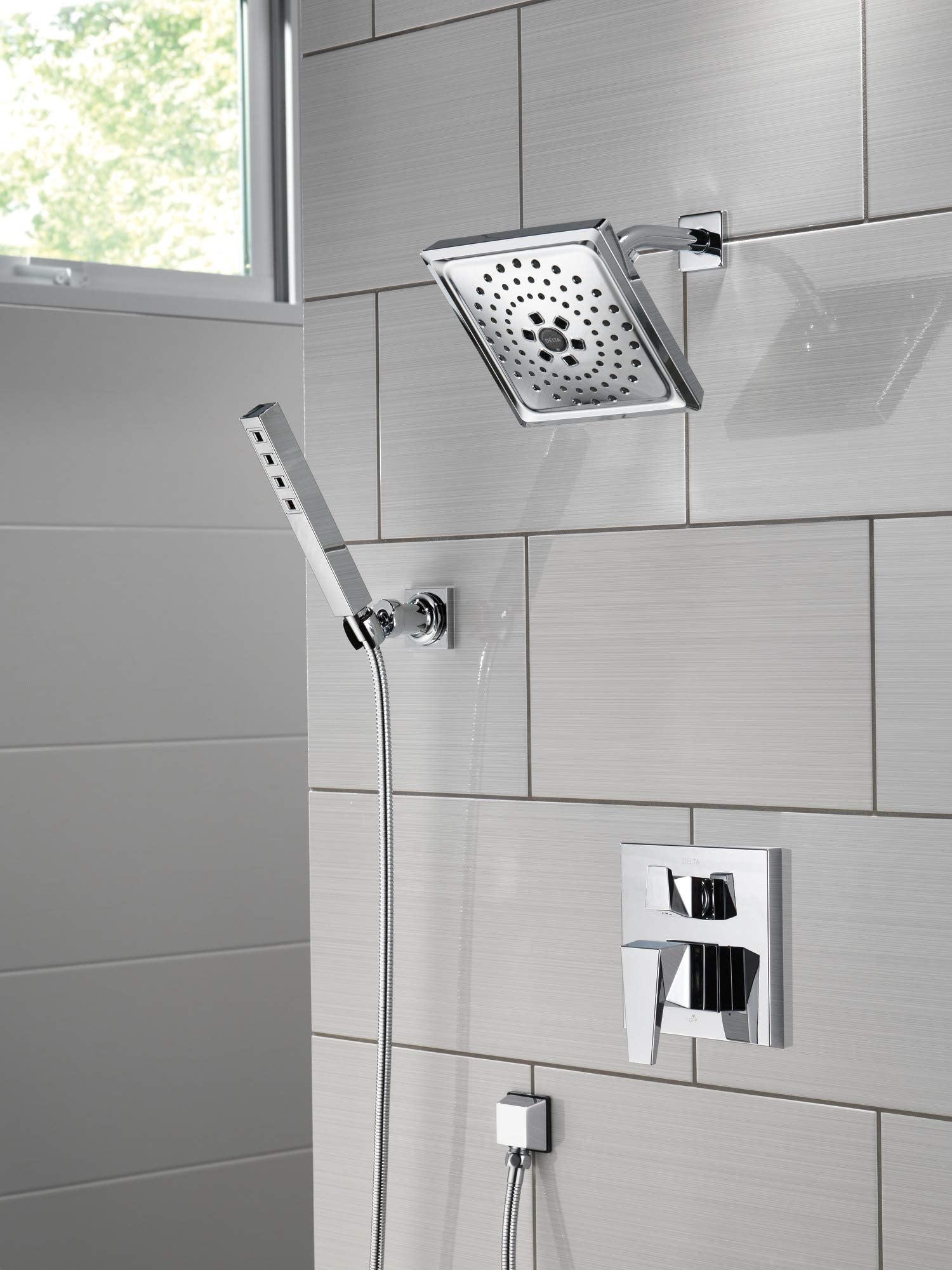 Square chrome shower head with perforated spray pattern mounted on tile wall with handheld shower wand and valve trim.