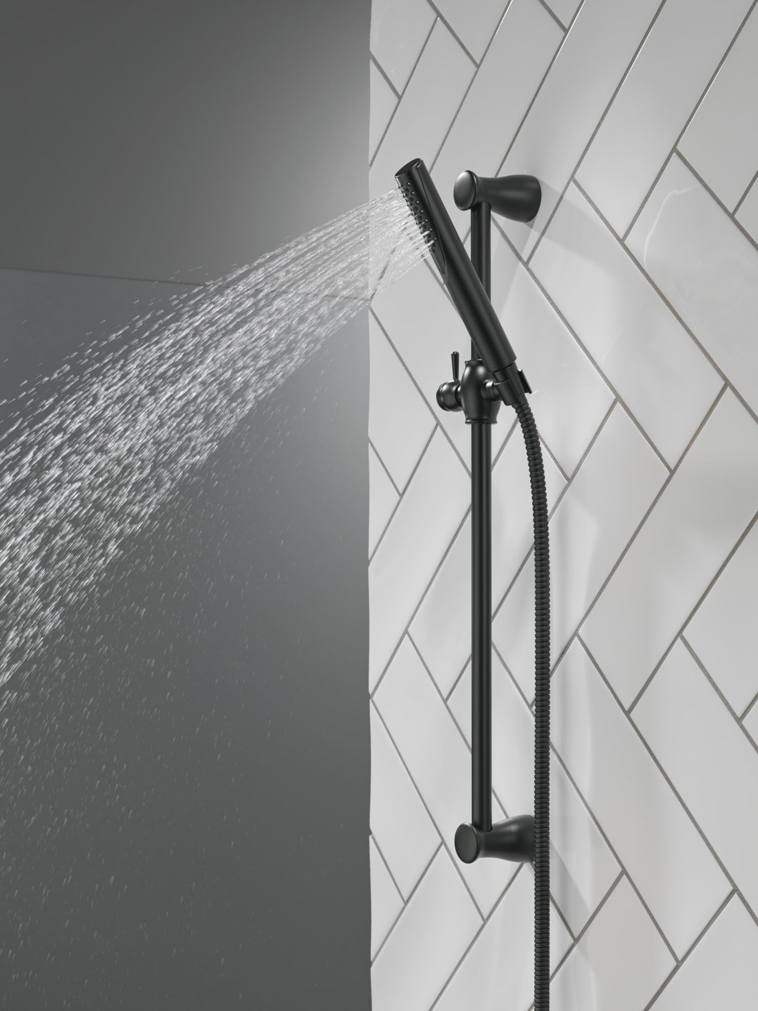 Matte black slide bar hand shower with water spray, mounted on white subway tile wall with hose connection.
