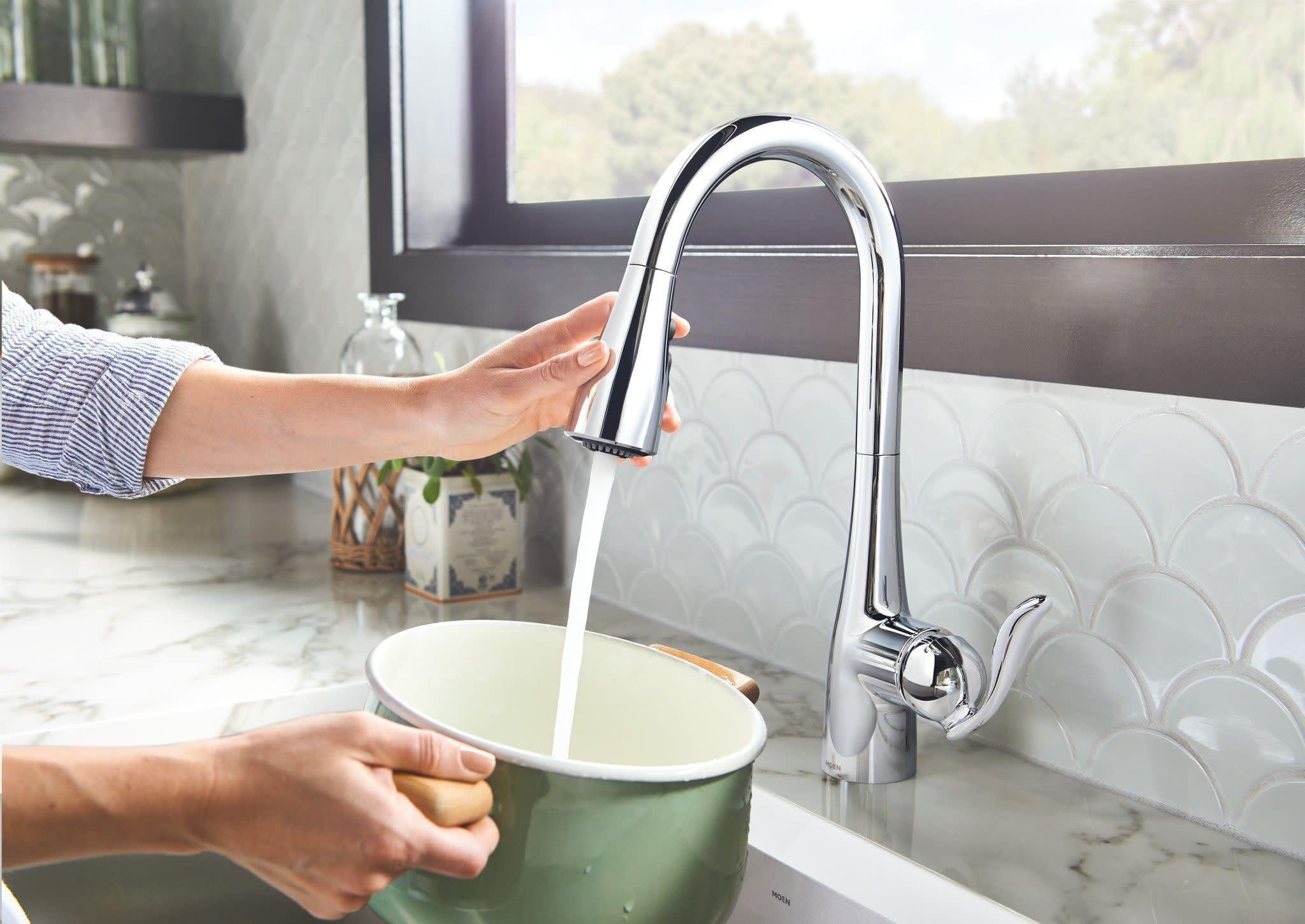 Chrome pulldown kitchen faucet with curved spout, installed above white marble sink with hand activating water stream.