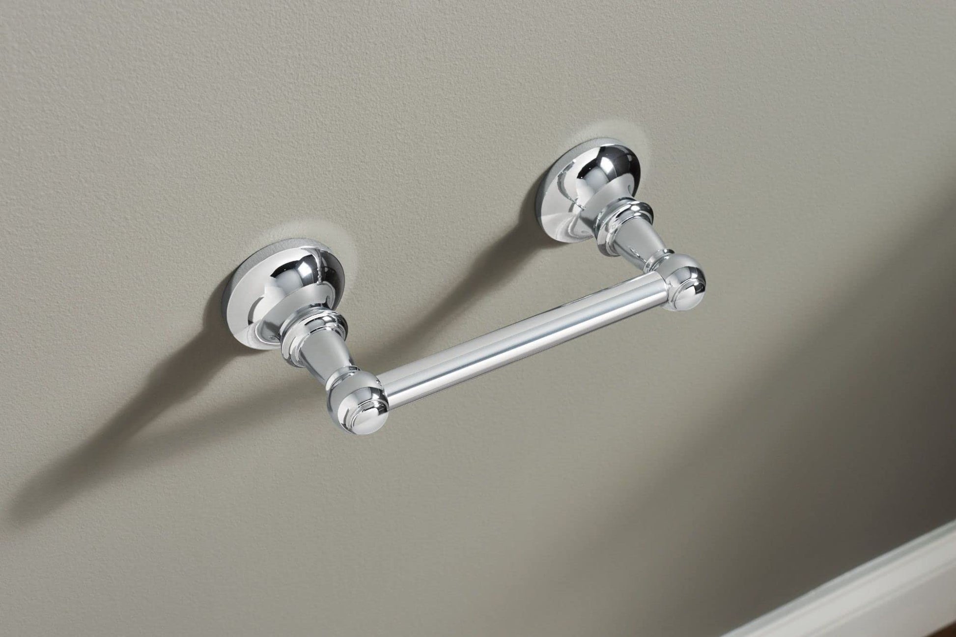 Chrome toilet paper holder with two circular wall-mounted brackets connected by a horizontal bar, installed on a gray wall.