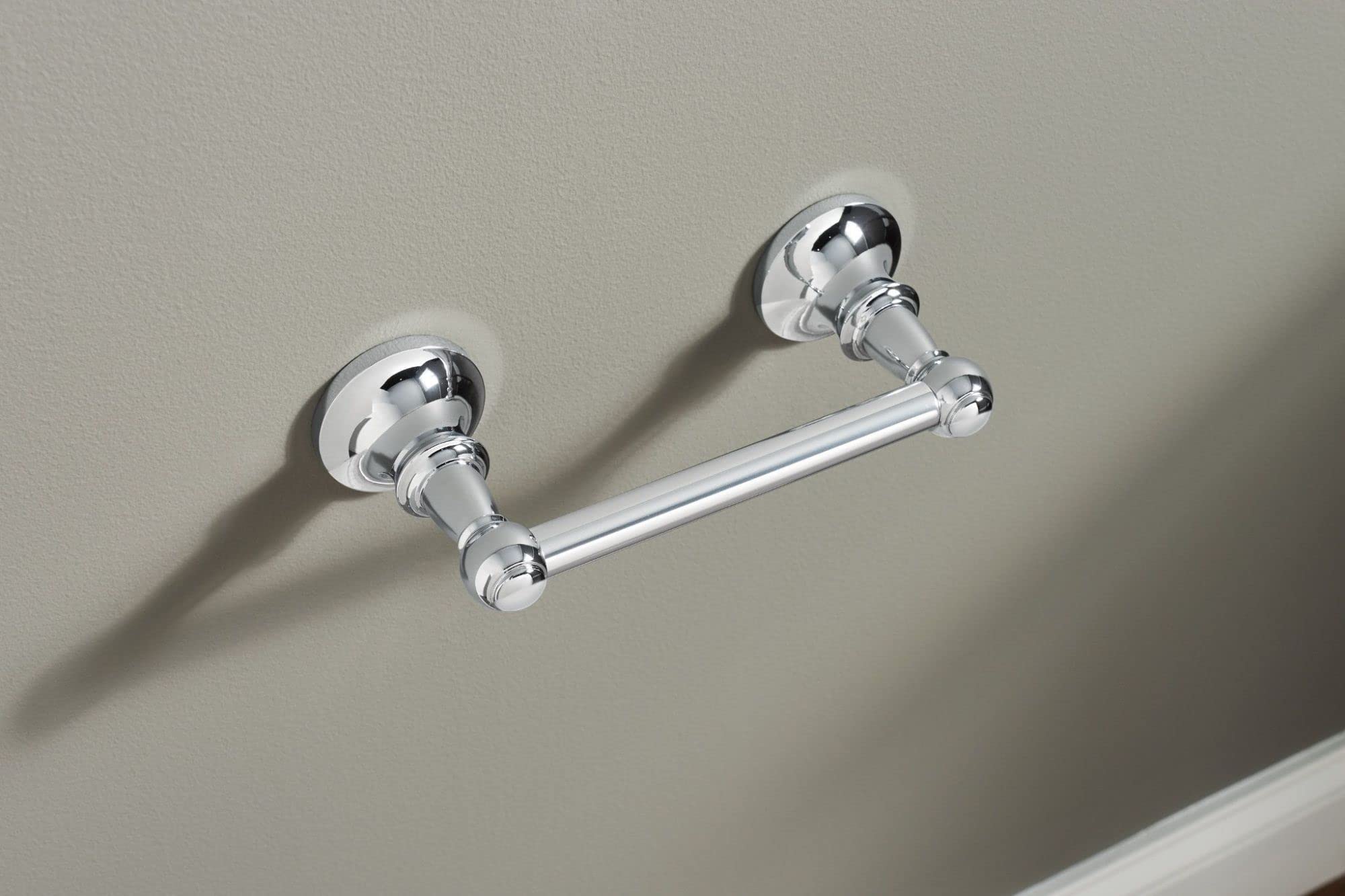 Chrome toilet paper holder with two circular wall-mounted brackets connected by a horizontal bar, installed on a gray wall.