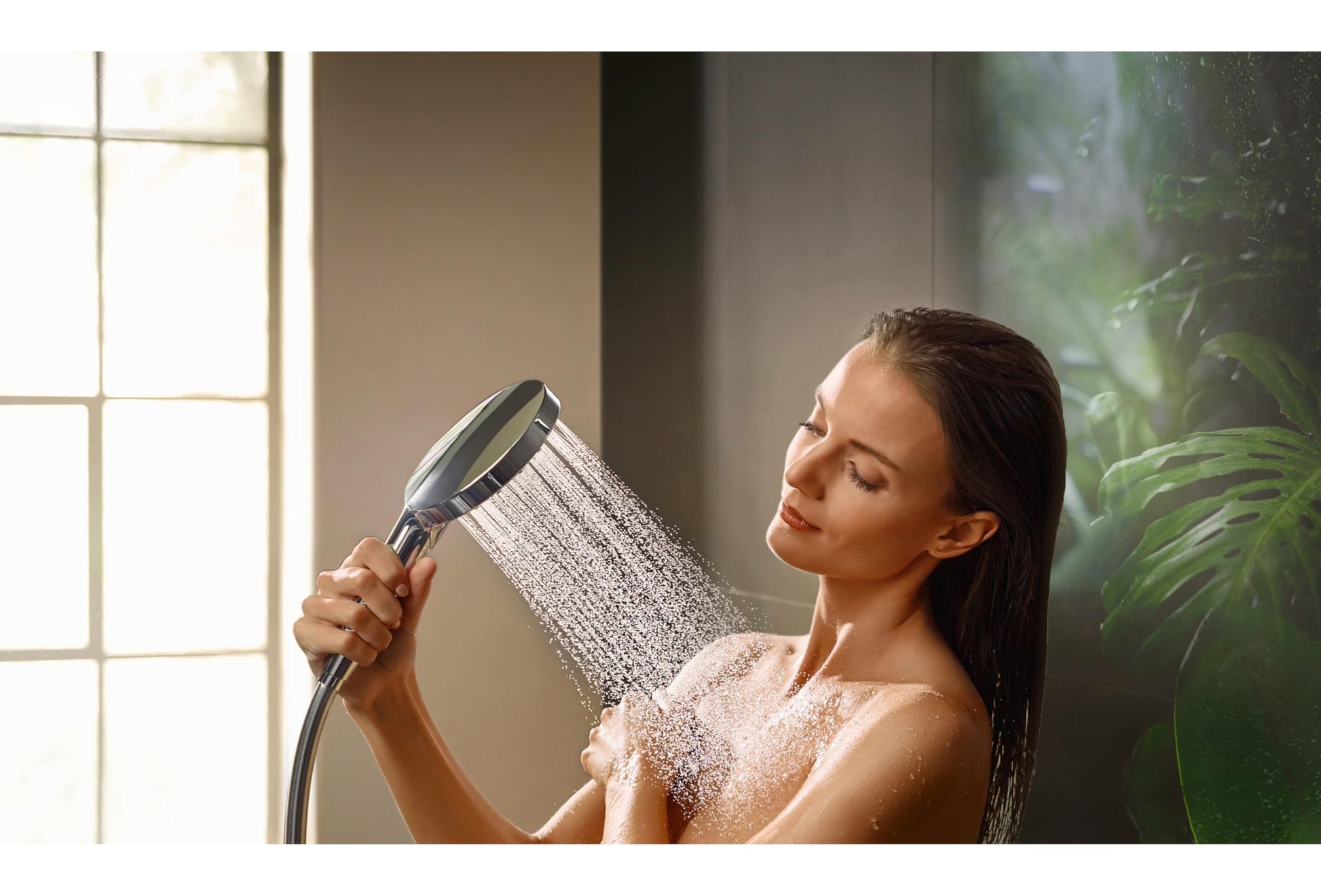 Chrome handheld showerhead with grip held by person under water spray in bathroom setting.