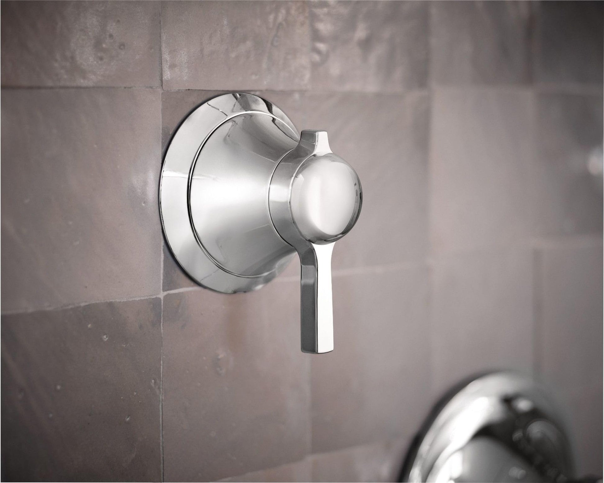 Chrome volume control valve trim with round escutcheon plate and lever handle mounted on tiled wall.
