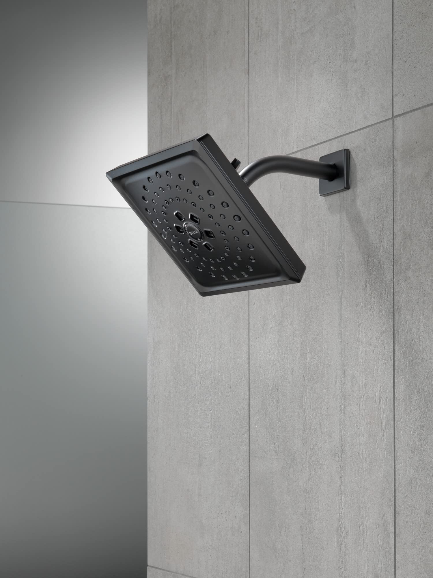 Matte black square shower head with spray holes mounted on curved wall arm against gray tile.