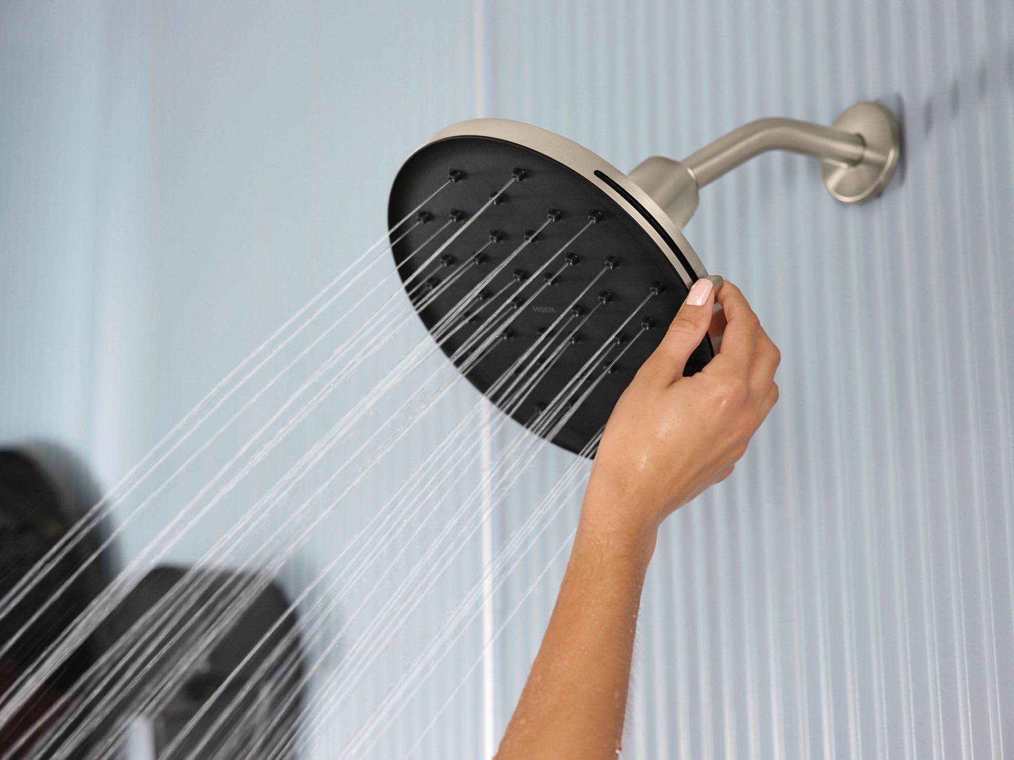 Brushed nickel round rainshower head mounted on wall with curved arm, dispensing water while hand adjusts spray pattern.