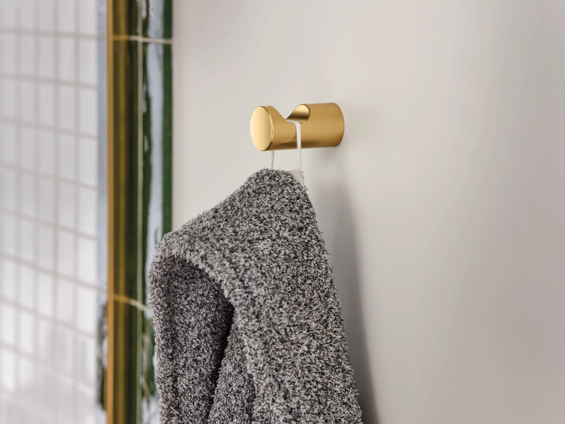 Brushed gold robe hook with cylindrical design mounted on white tile wall, holding a gray towel.
