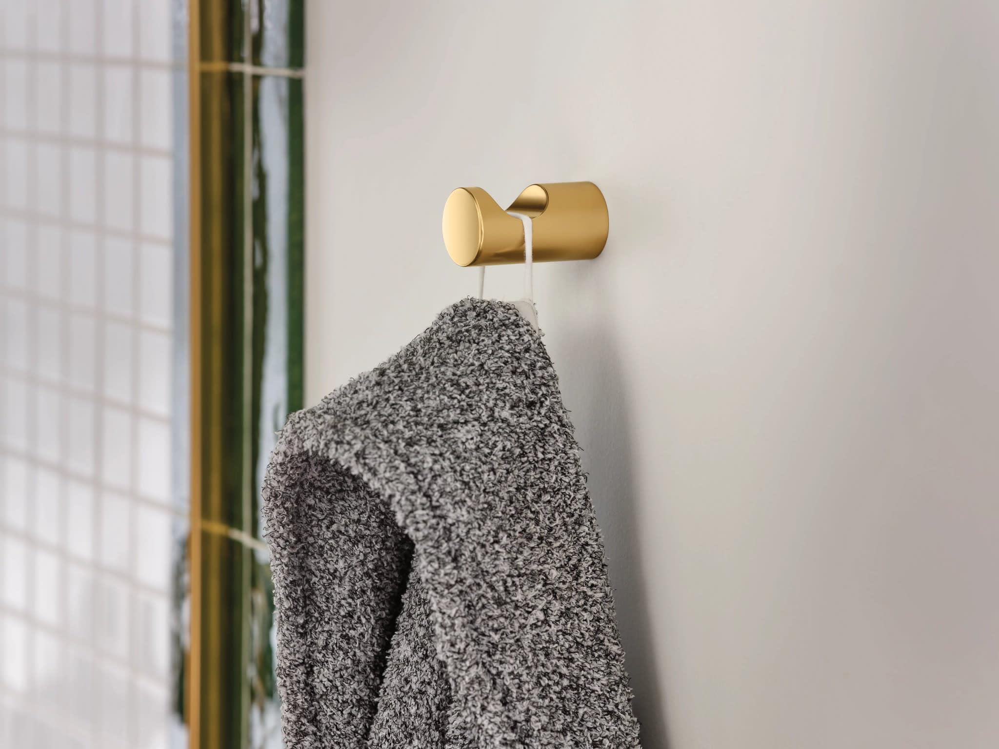 Brushed gold robe hook with cylindrical design mounted on white tile wall, holding a gray towel.