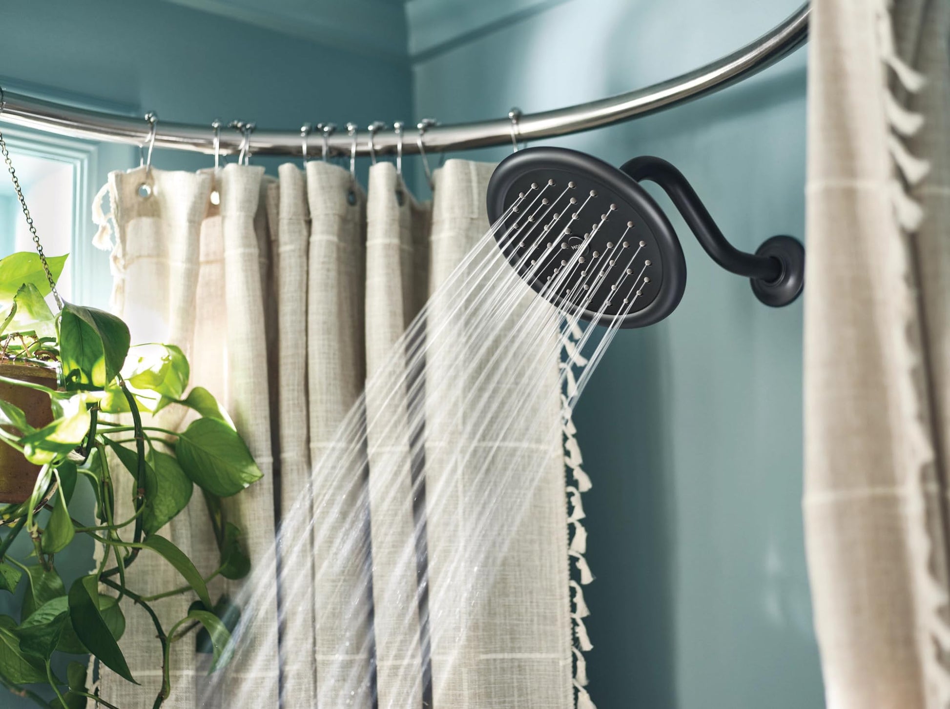 Matte black oval showerhead mounted on shower wall with water streaming down, curved chrome shower rod and cream curtain visi