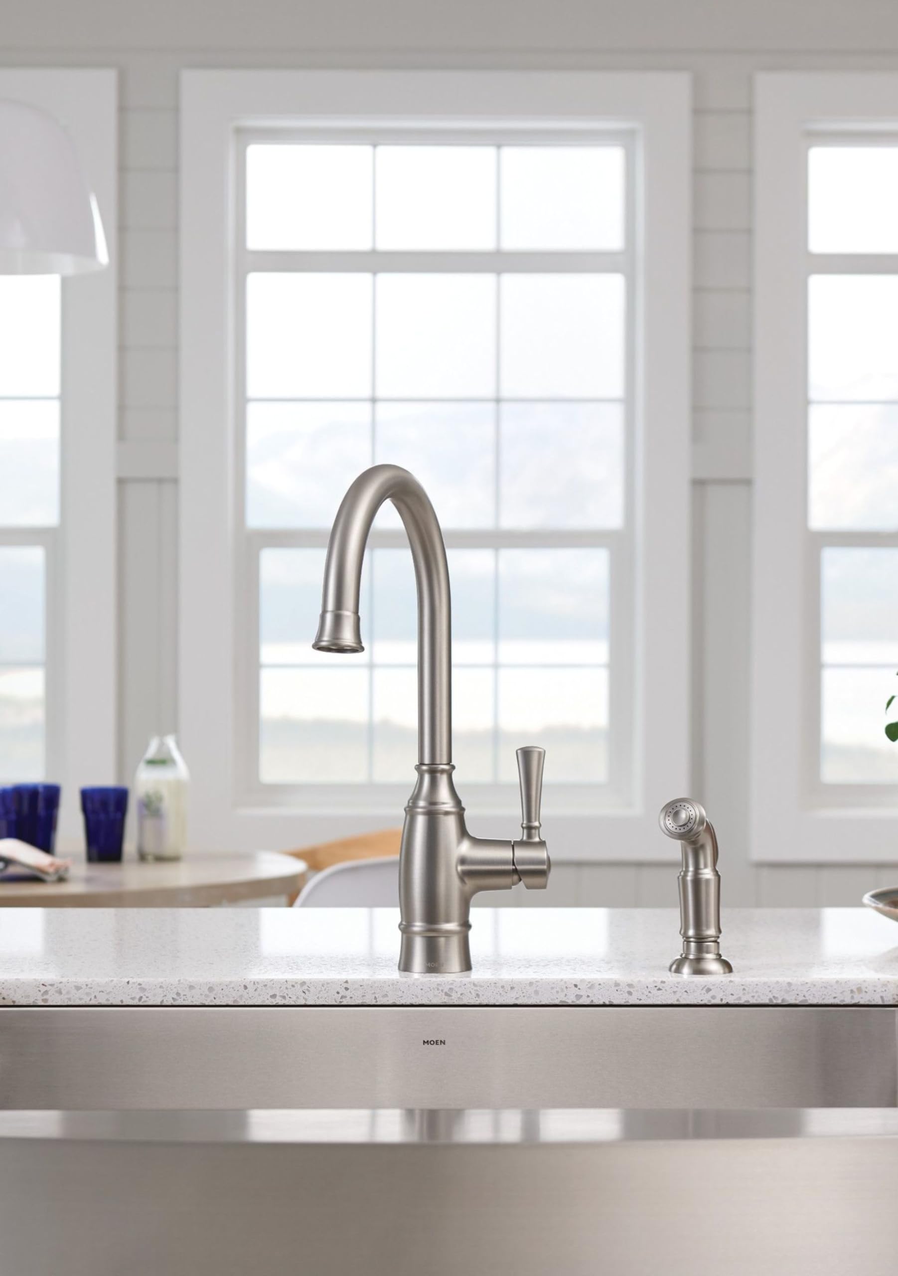 Stainless steel farmhouse apron front kitchen sink with curved faucet and side sprayer installed in white countertop.