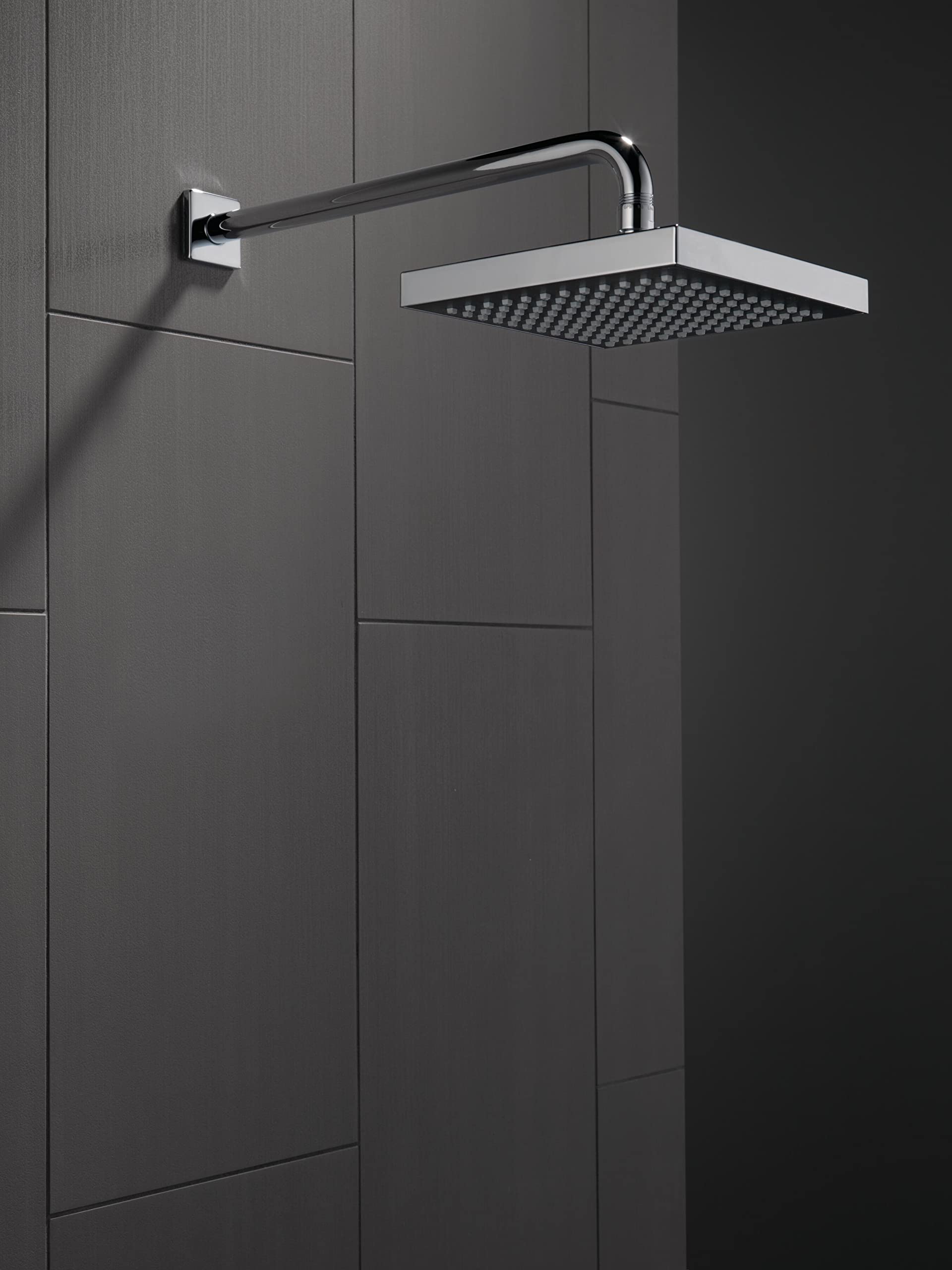 Chrome raincan shower head with square face mounted on curved wall arm against dark tile wall.