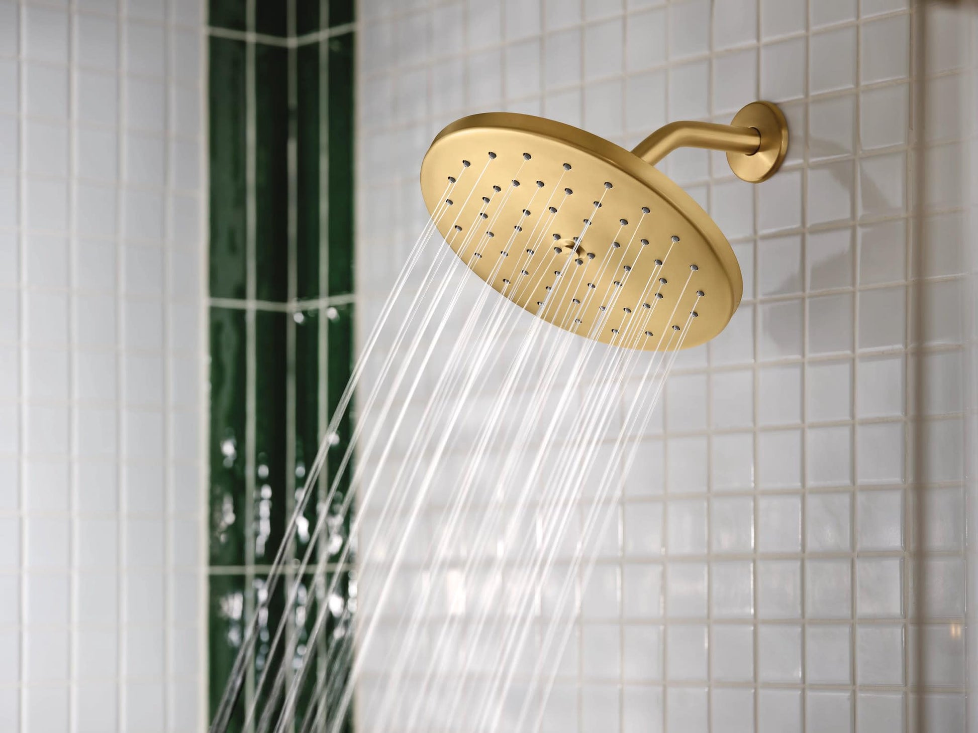Brushed gold round showerhead with water streams flowing, mounted on tile wall with green plant visible.