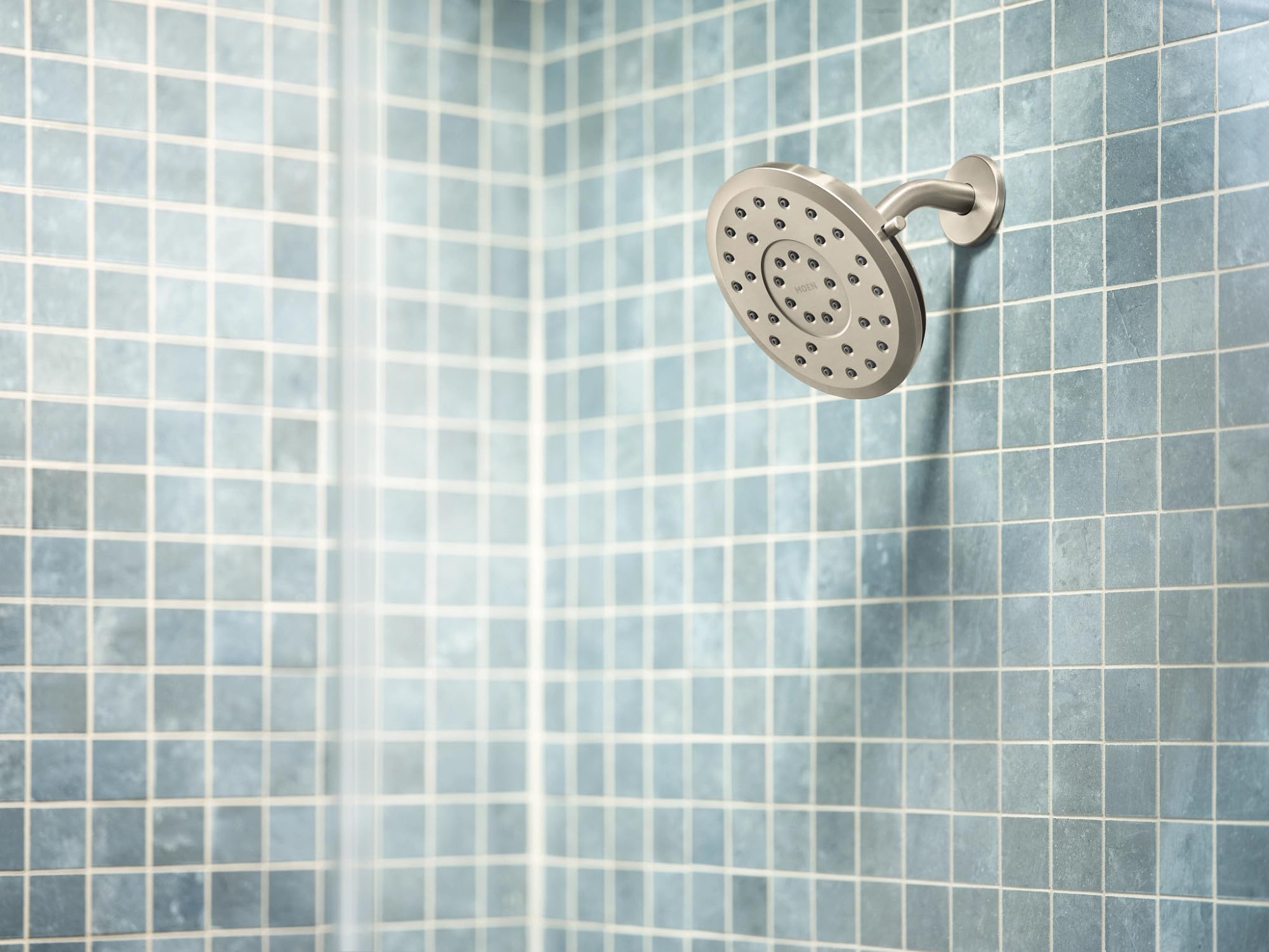 Round brushed nickel rain shower head with multiple spray holes mounted on tile wall with curved arm.