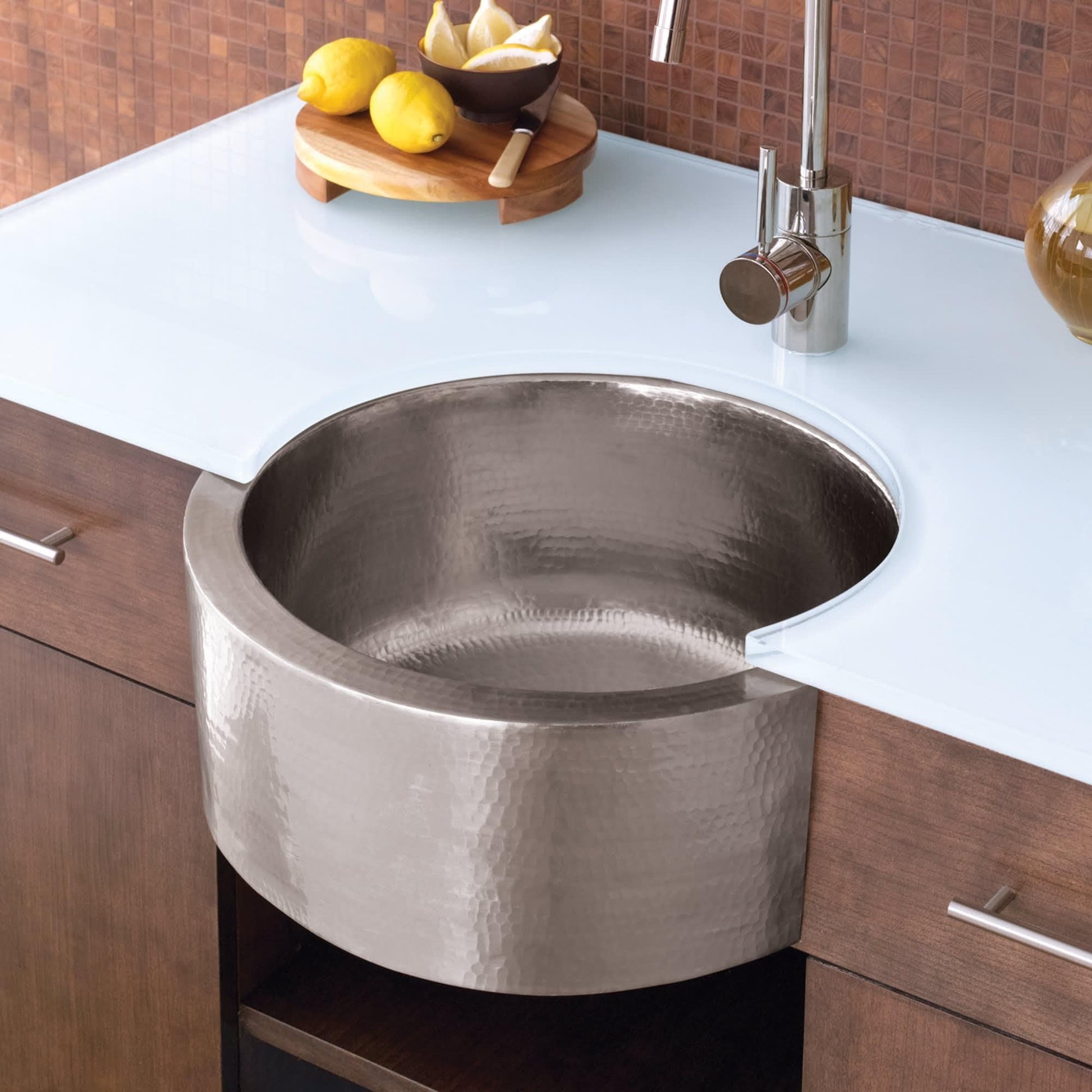 Hammered copper undermount sink with round basin installed in white countertop with chrome faucet and wood cabinetry.