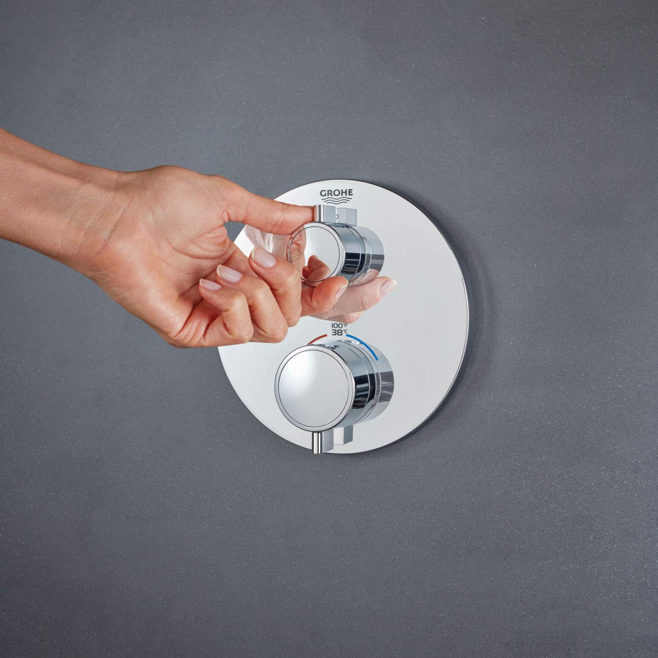Chrome thermostatic valve trim with two handles mounted on white circular plate, hand adjusting upper control.