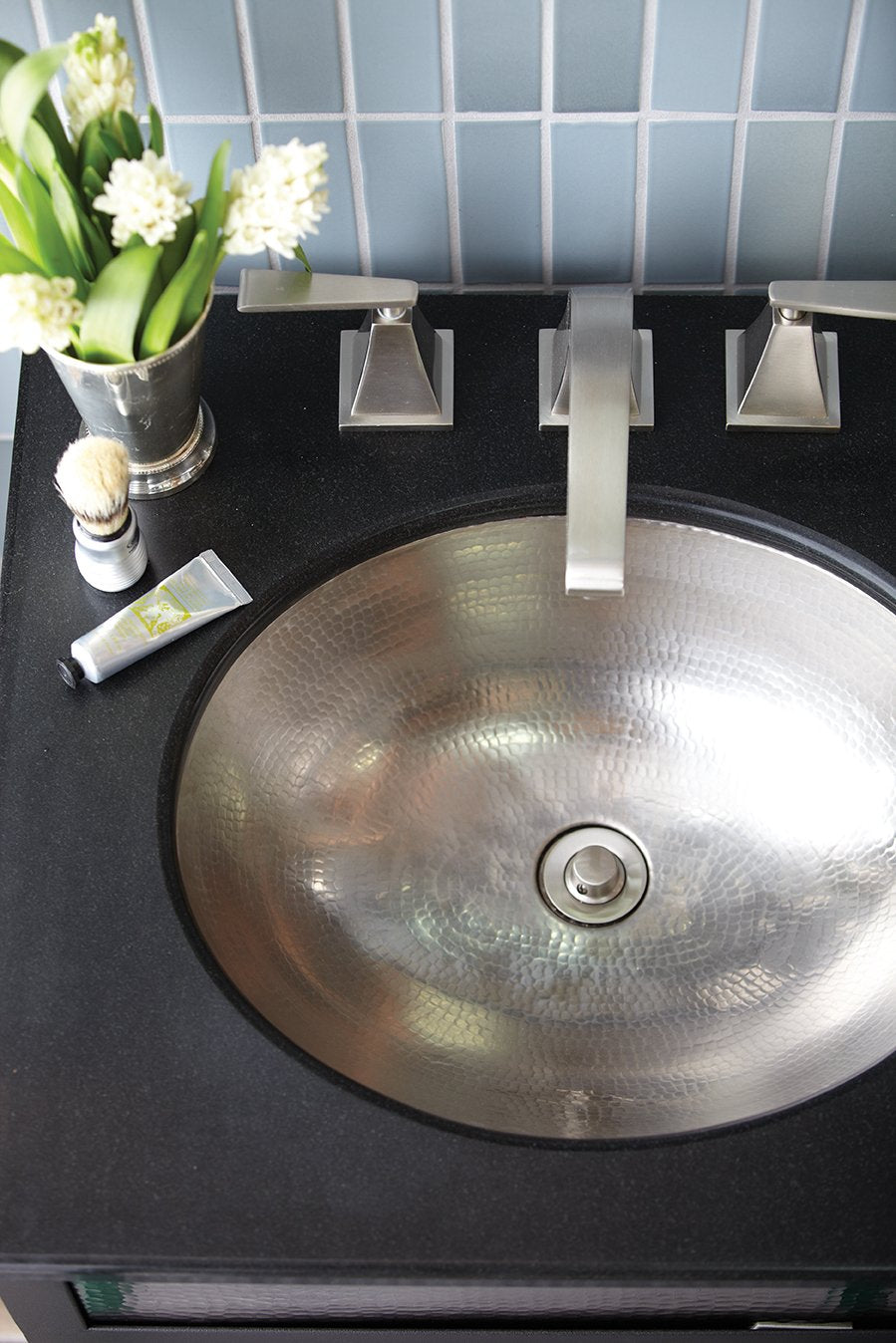 Hammered copper round bathroom sink with brushed nickel faucet and handles installed on black countertop.