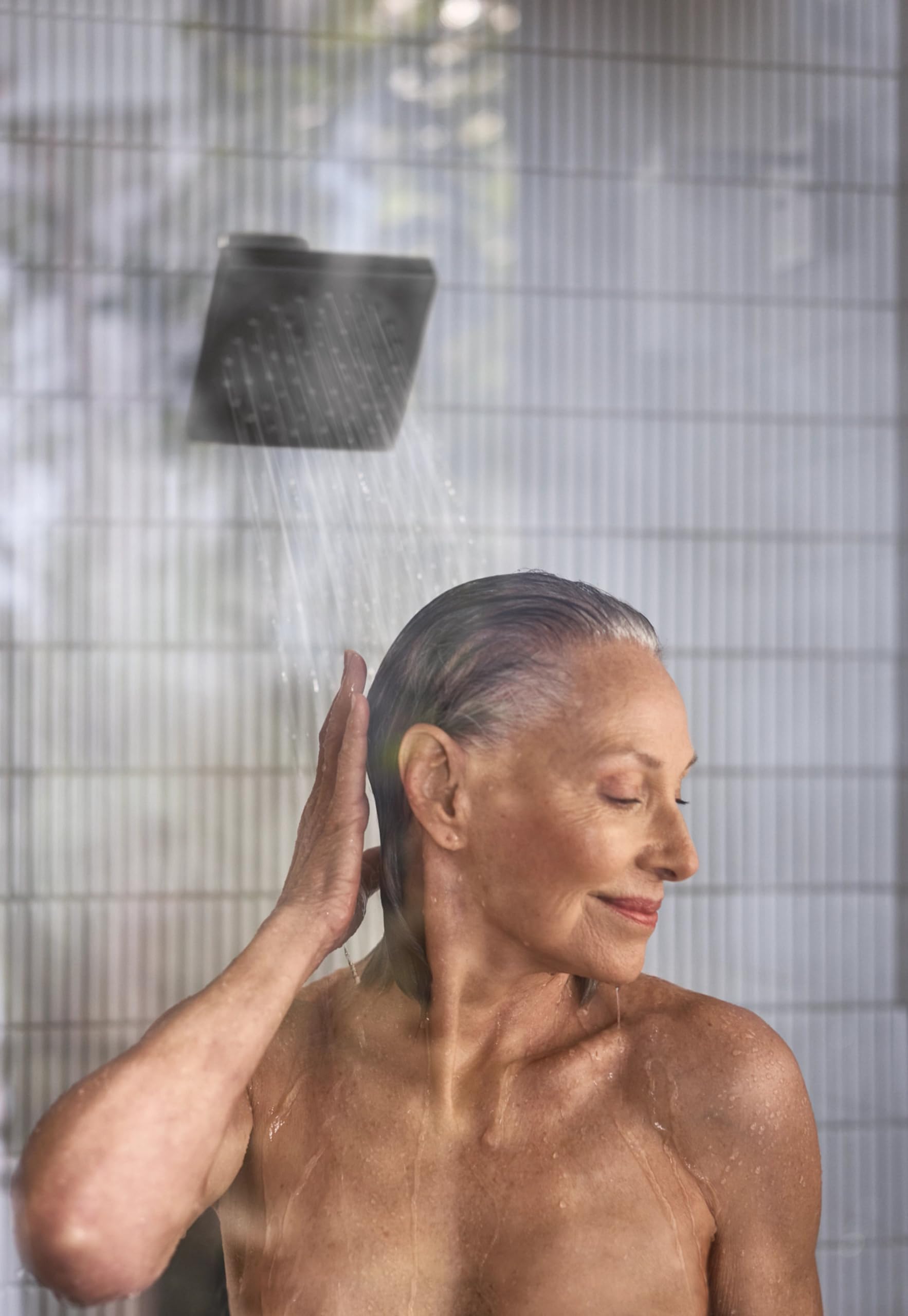 Matte black square shower head with water stream spraying over woman's head in tiled shower enclosure.