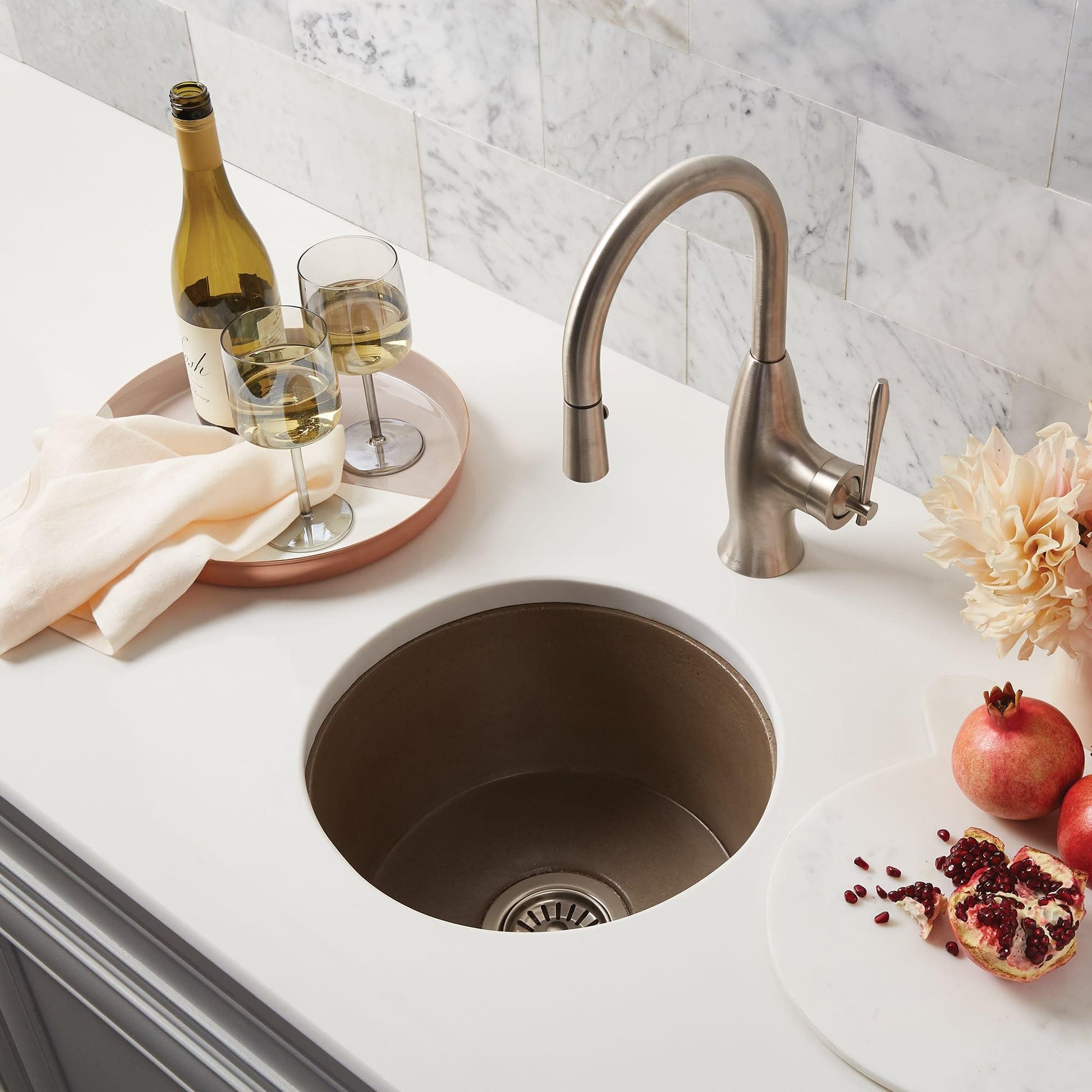Slate bar sink with curved stainless steel faucet, drain strainer, and white countertop decorated with wine glasses and fruit