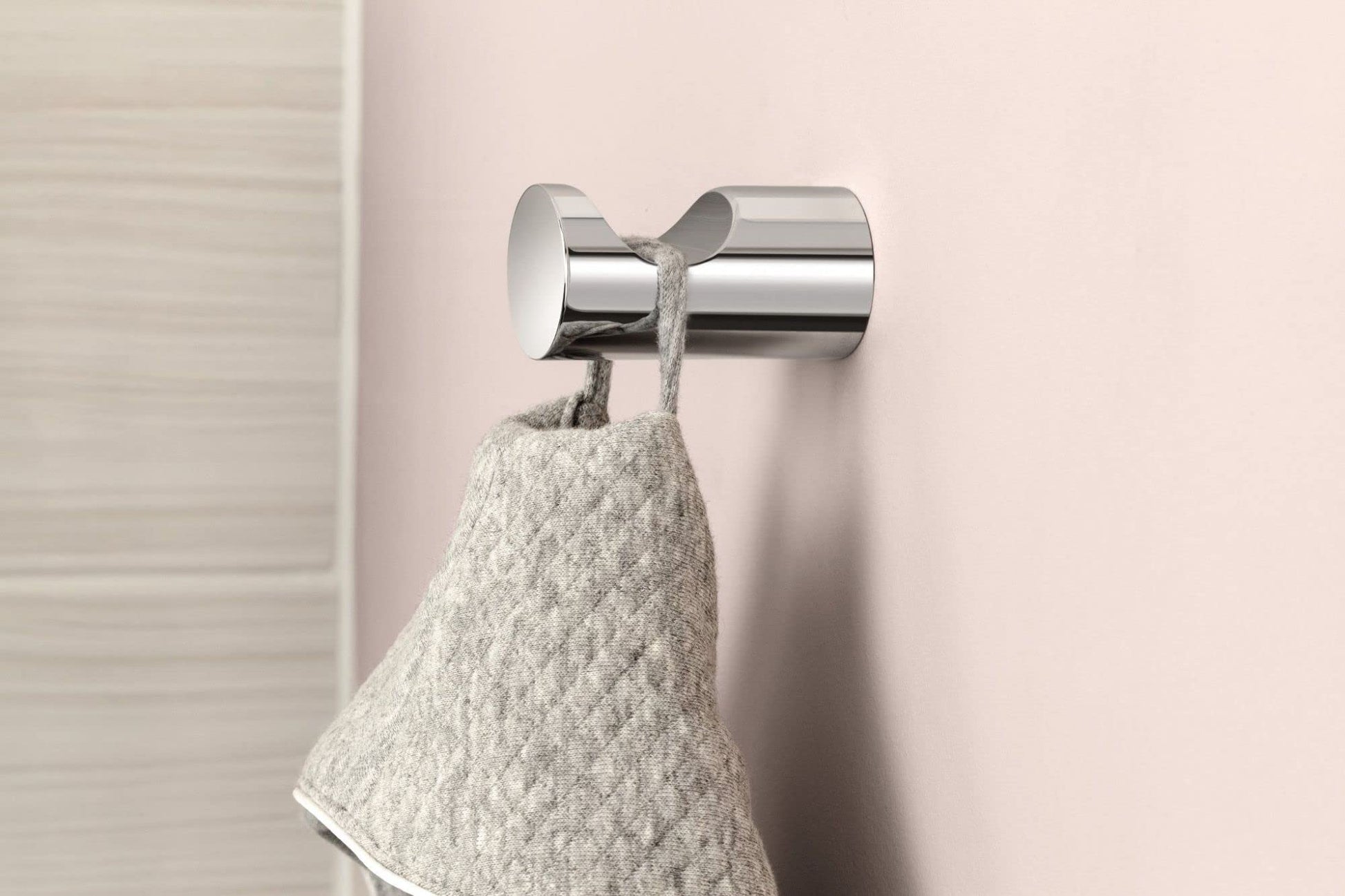 Chrome robe hook with cylindrical design holding a light gray towel against a pink wall.