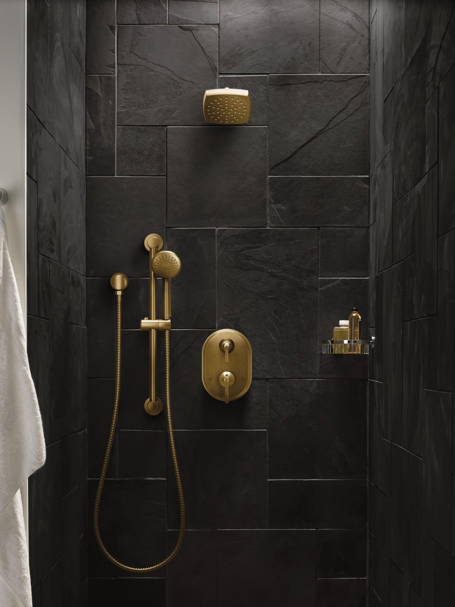 Brushed gold hand shower with perforated face mounted on slide bar above dark tile shower wall.