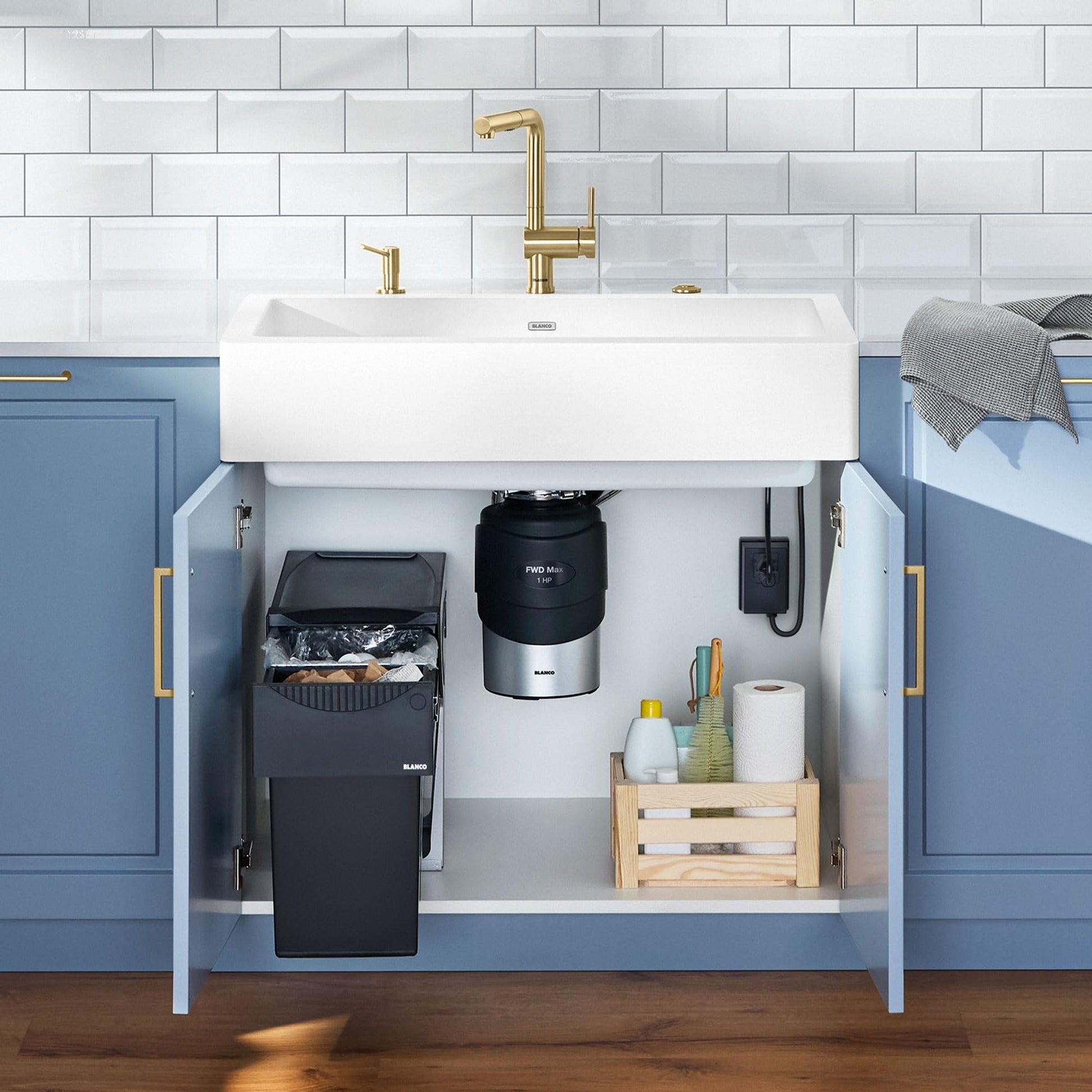 Black Blanco food waste disposal unit installed under white sink with brass faucet and blue cabinets.