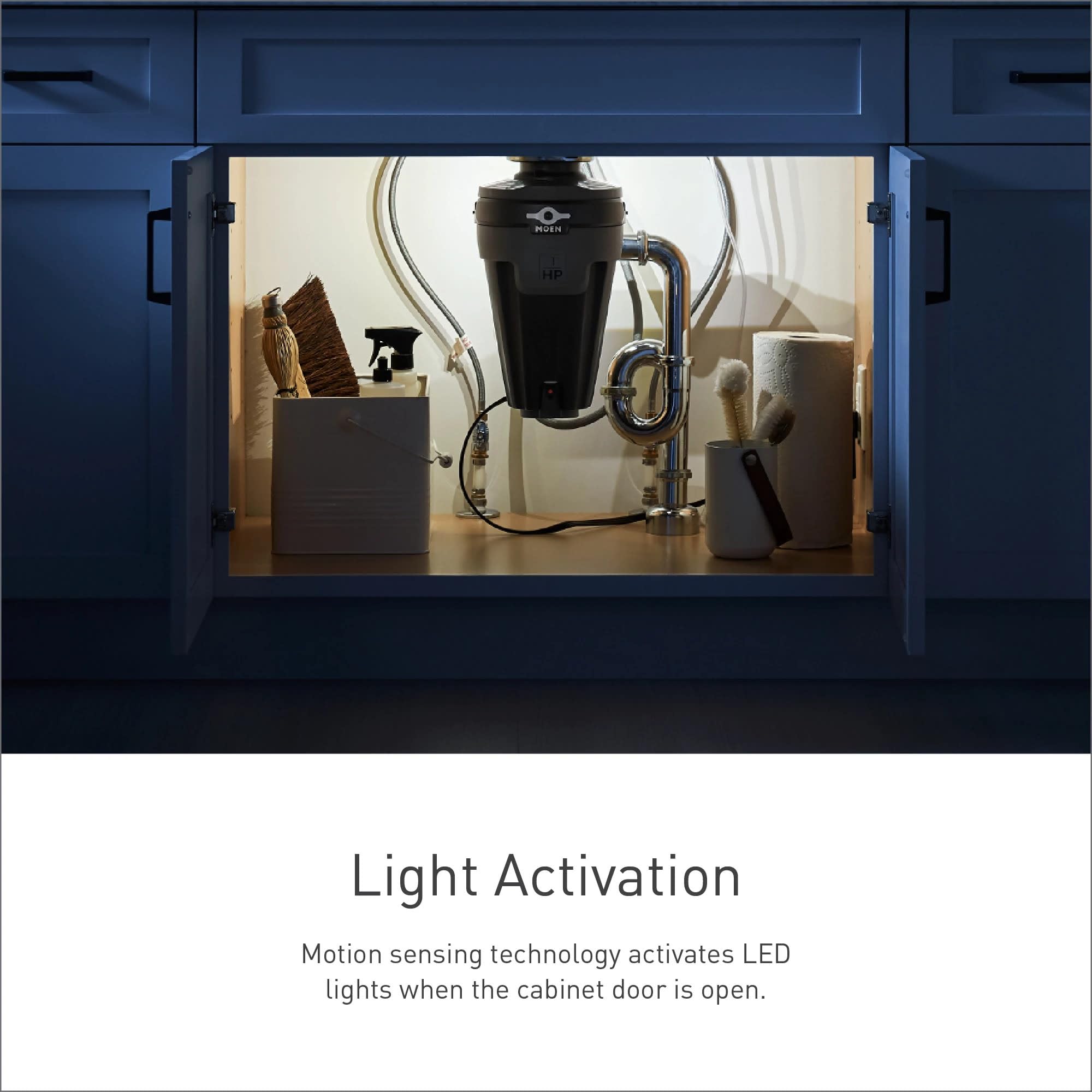 Black garbage disposal unit installed under kitchen sink with chrome pipes, LED light illuminated inside open cabinet.