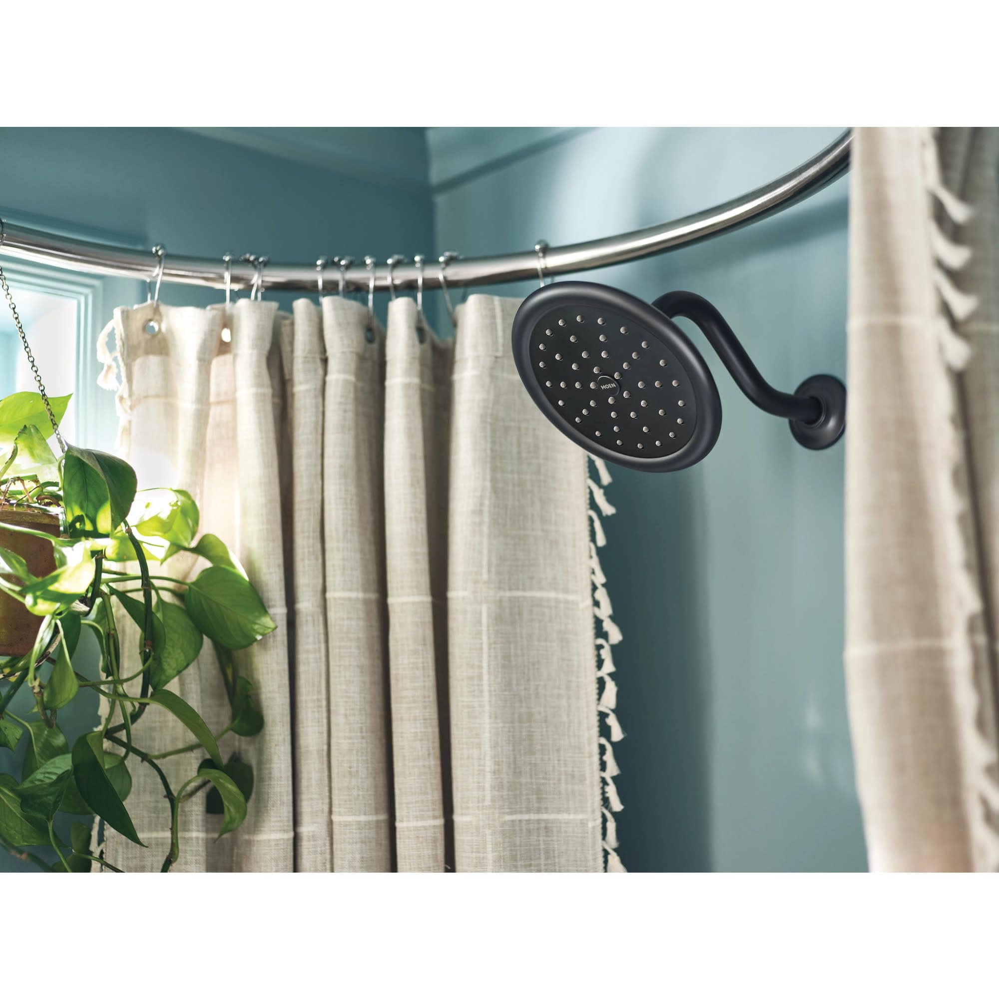 Oil-rubbed bronze shower head and trim mounted on tiled bathroom wall with curved shower rod and cream curtain.