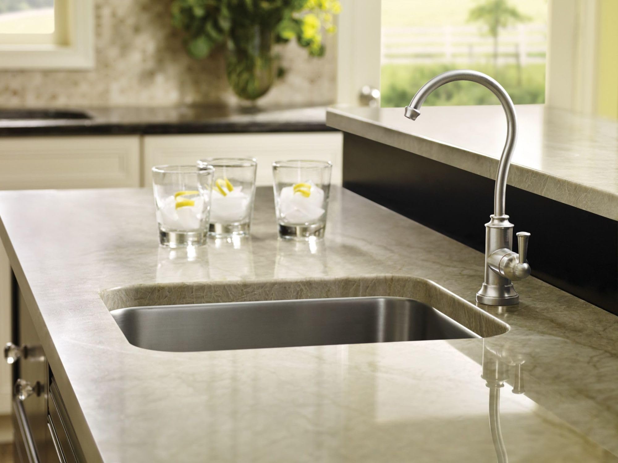 Stainless steel kitchen faucet with curved spout mounted on countertop sink next to glasses of water with lemon.