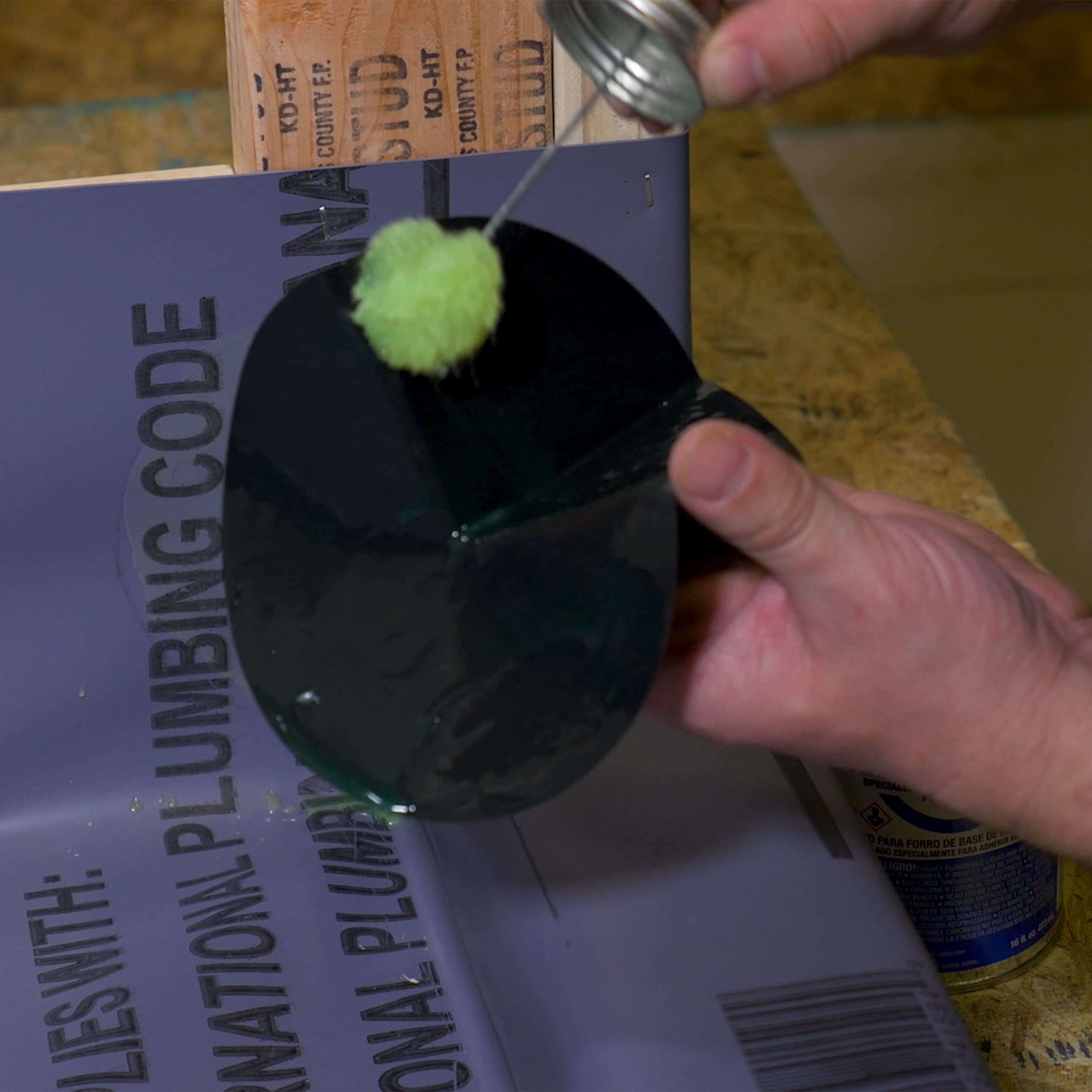 Hand holding dark-capped PVC solvent cement container with brush applicator over product packaging.