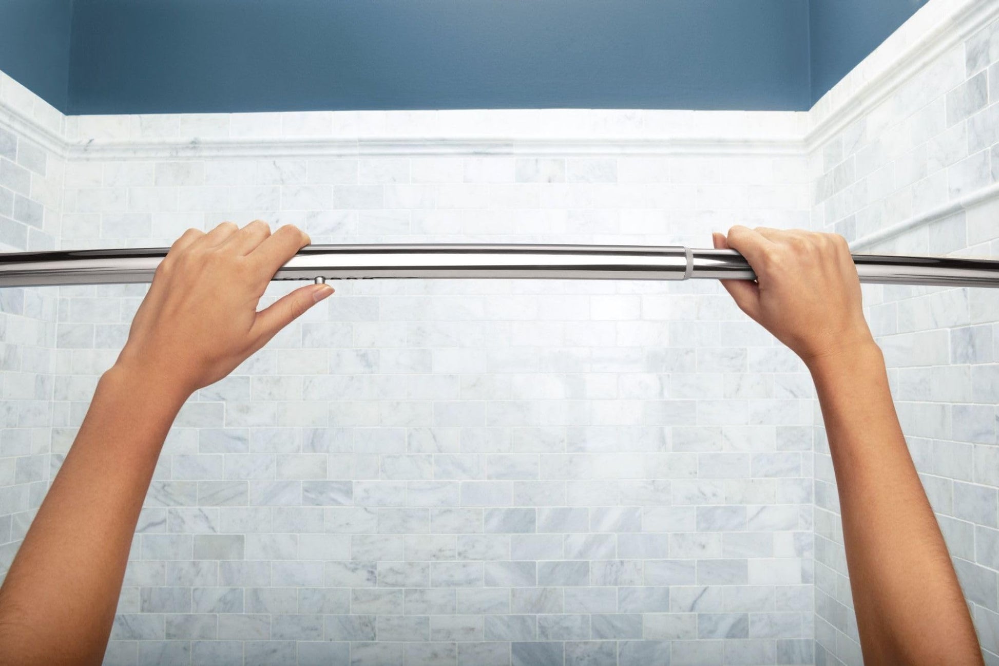 Curved chrome shower rod installed between two tiled shower walls with hands adjusting the rod.
