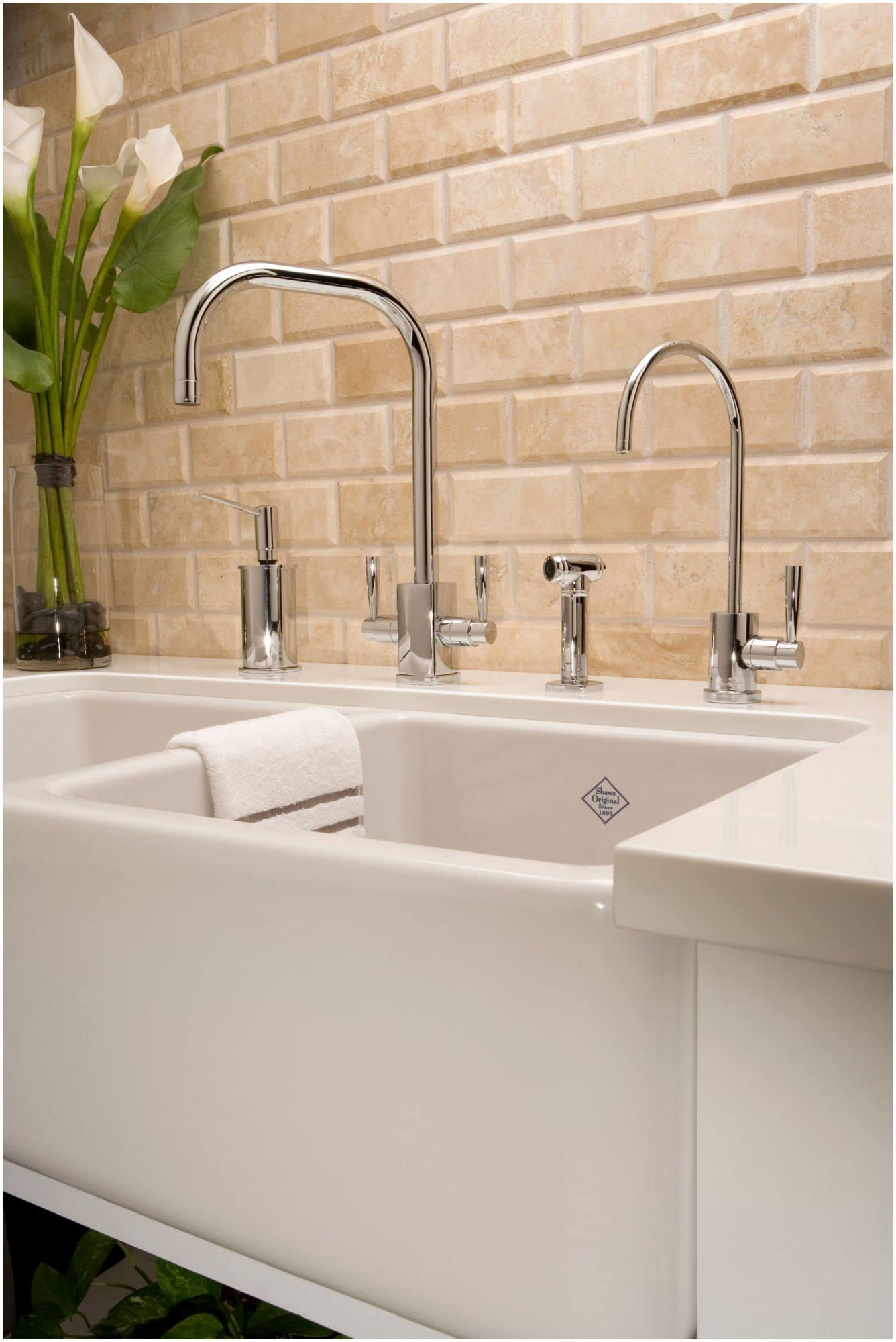 Polished chrome kitchen faucets with curved spouts installed on white farmhouse sink against tan brick backsplash.