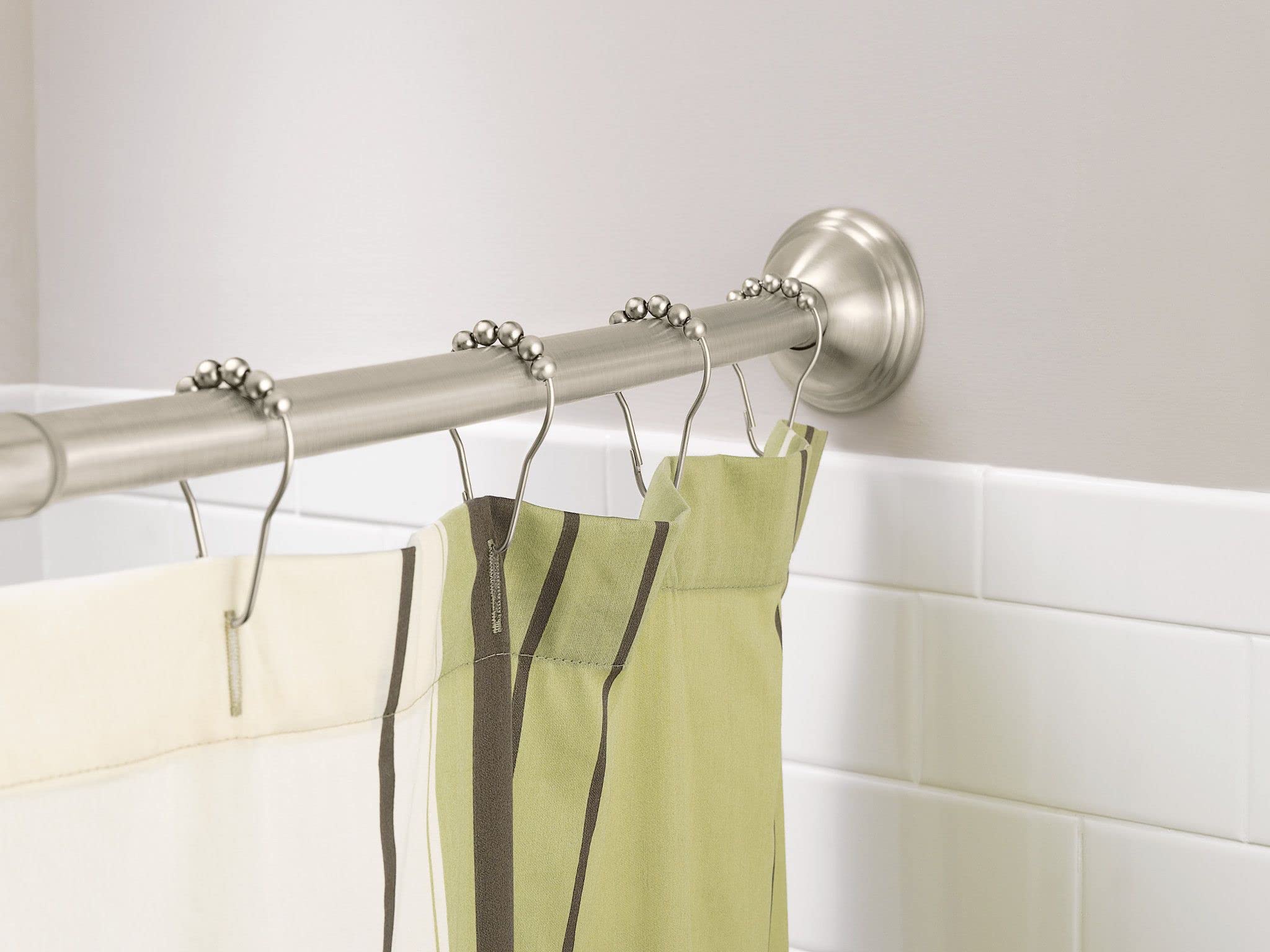Brushed nickel curved shower rod with decorative rings holding green and cream curtains in a white tiled bathroom.