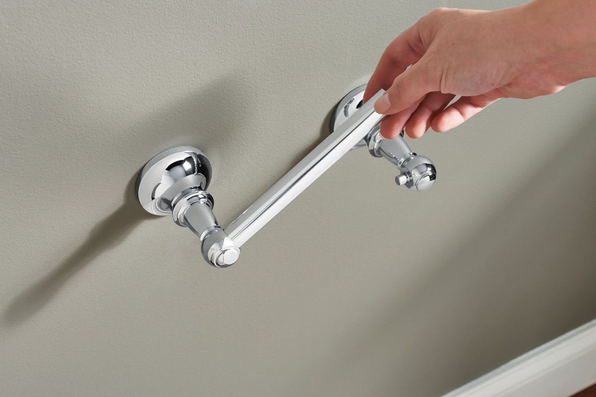 Chrome toilet paper holder mounted on wall with cylindrical bar and circular mounting flanges, shown with hand demonstrating 