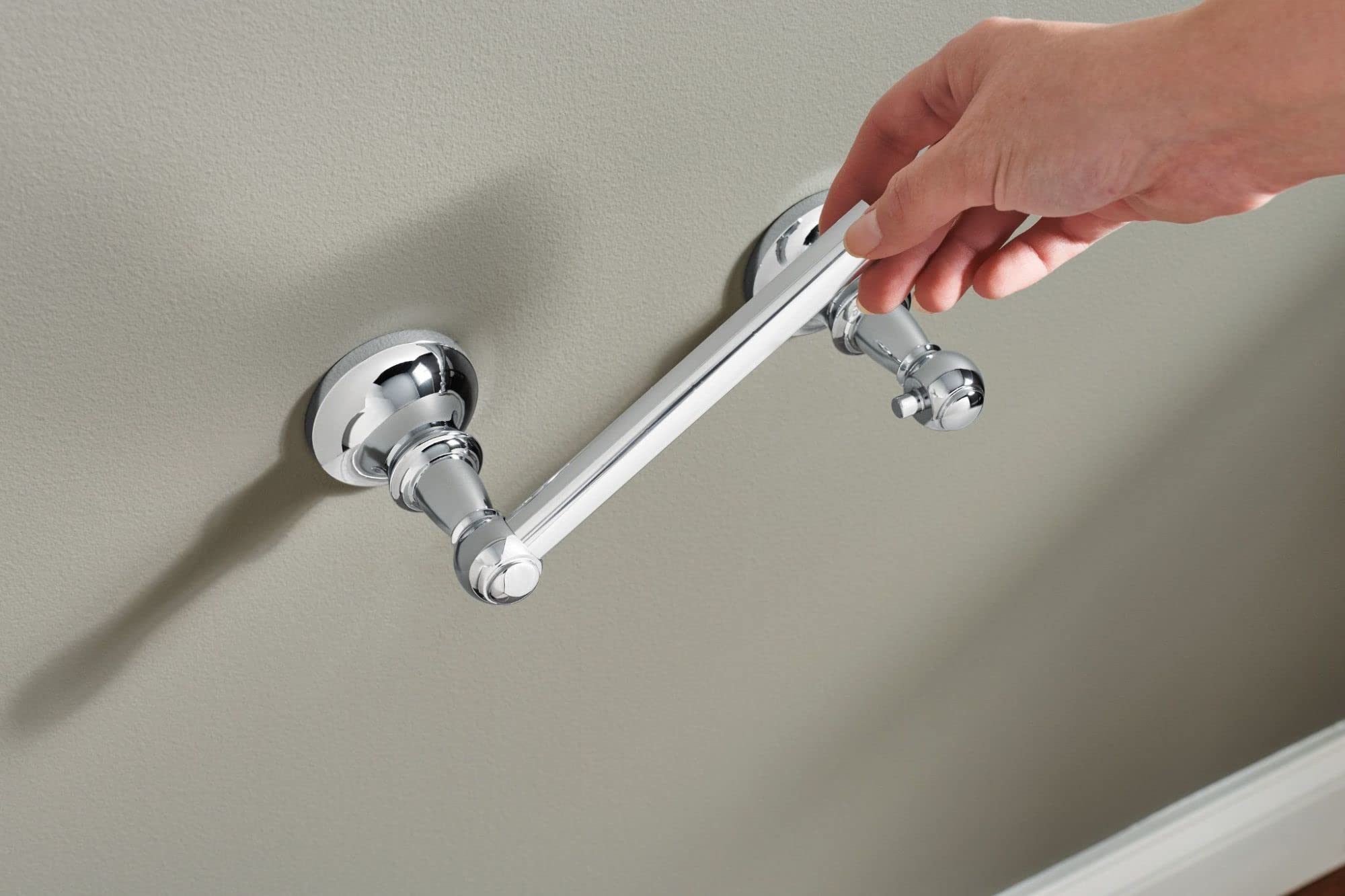 Chrome toilet paper holder mounted on wall with cylindrical bar and circular mounting flanges, shown with hand demonstrating 