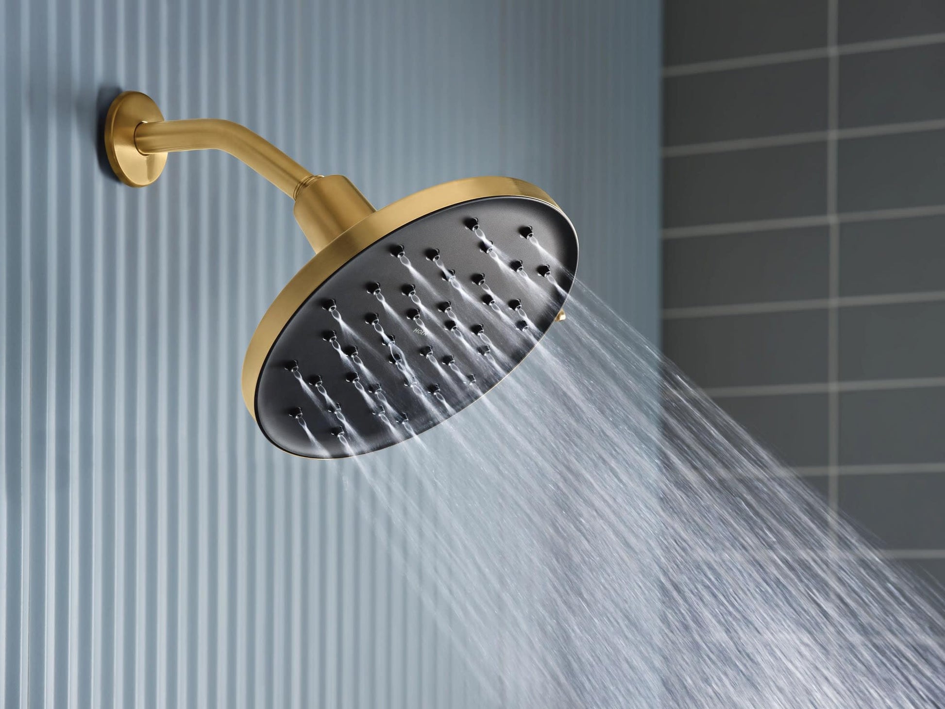 Brushed gold curved arm with round black rainshower head spraying water against tile wall background.
