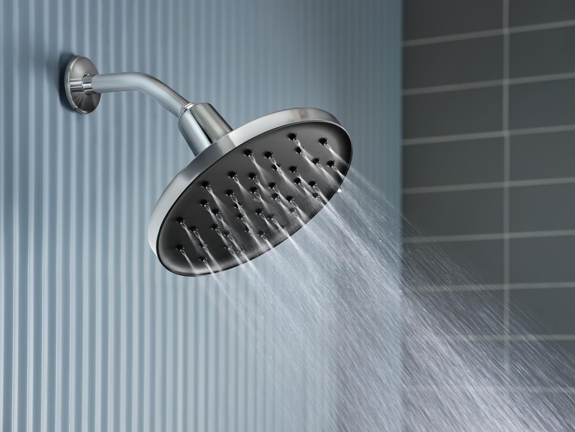 Chrome rainshower showerhead with round spray face mounted on angled arm against tiled bathroom wall.