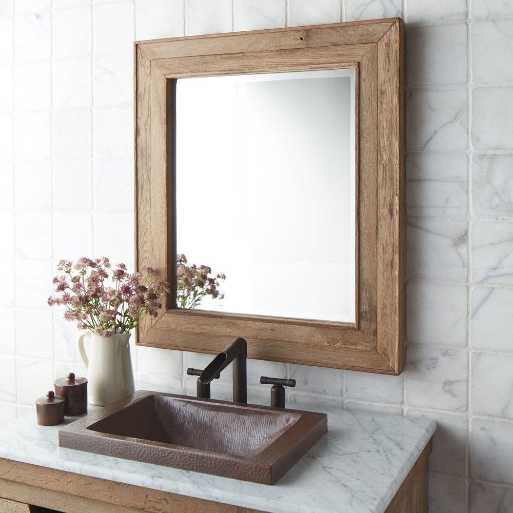 Rectangular mirror with light wood frame mounted above bathroom sink with black faucet and marble countertop.
