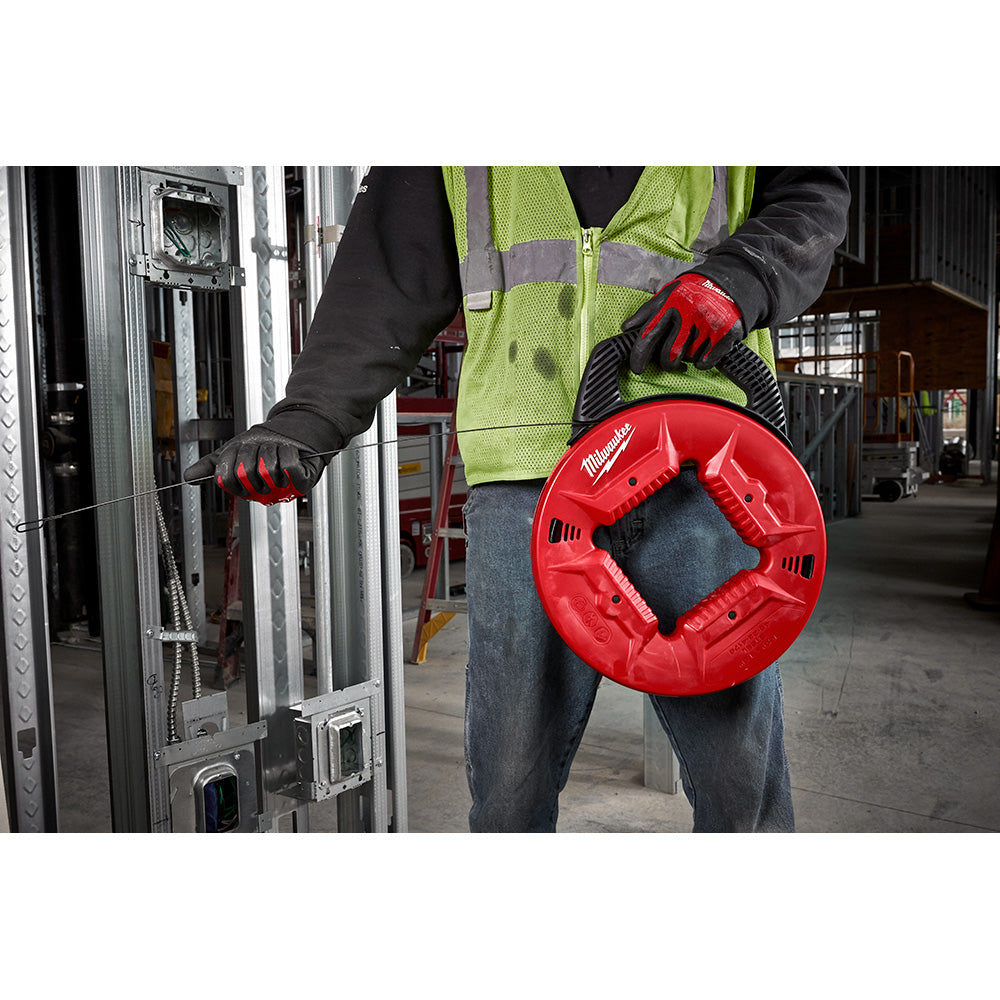 Worker in safety gear holding red Milwaukee fish tape reel while feeding steel cable into wall conduit.