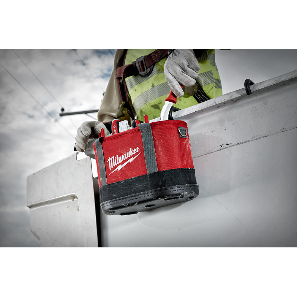 Red and black Milwaukee oval tool bag with gray straps held by worker on ladder.