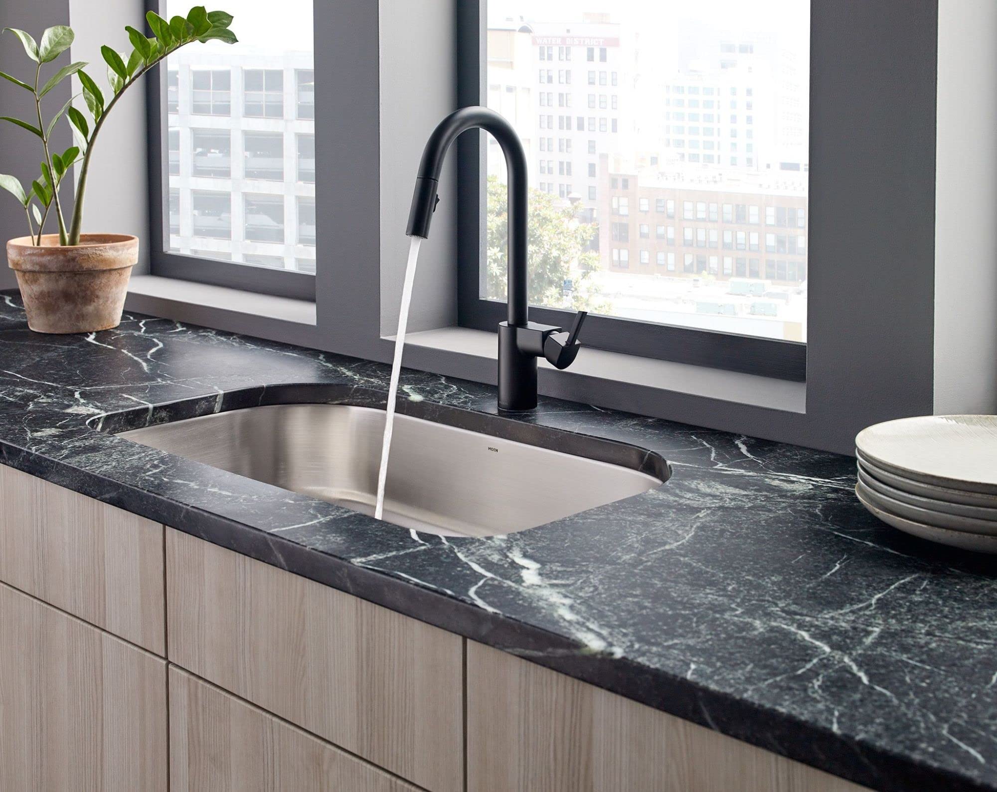 Matte black pulldown kitchen faucet with curved spout installed above stainless steel sink on dark marble countertop.