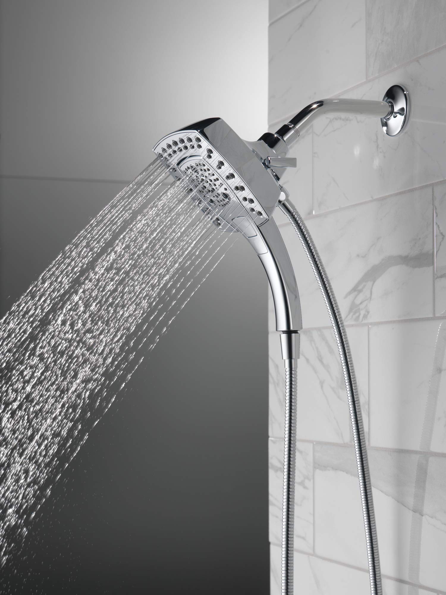 Chrome two-in-one shower head with handheld spray attached to curved arm and flexible hose against marble tile wall.