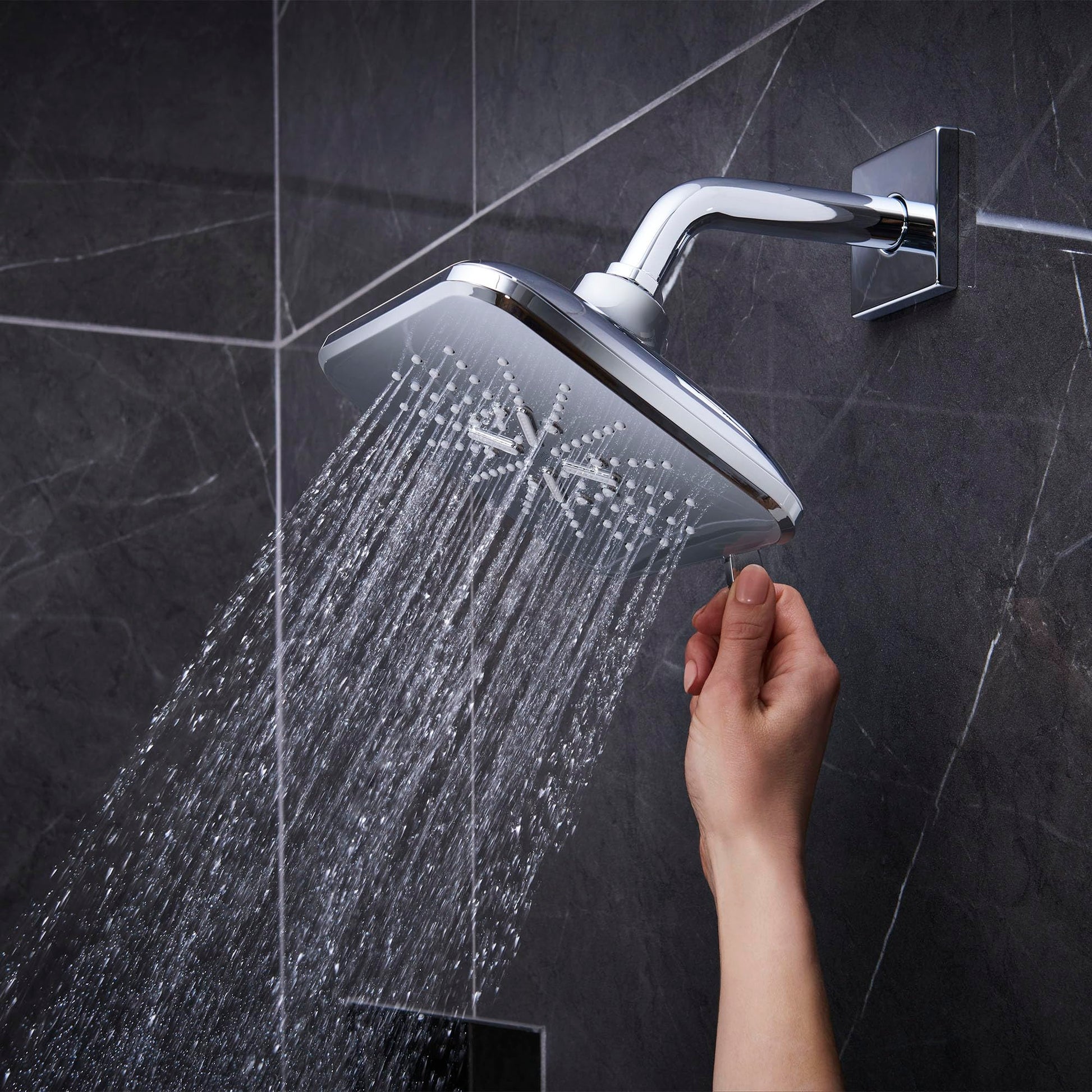 Chrome shower head with curved arm mounted on dark tile wall, dispensing water with hand reaching toward controls below.