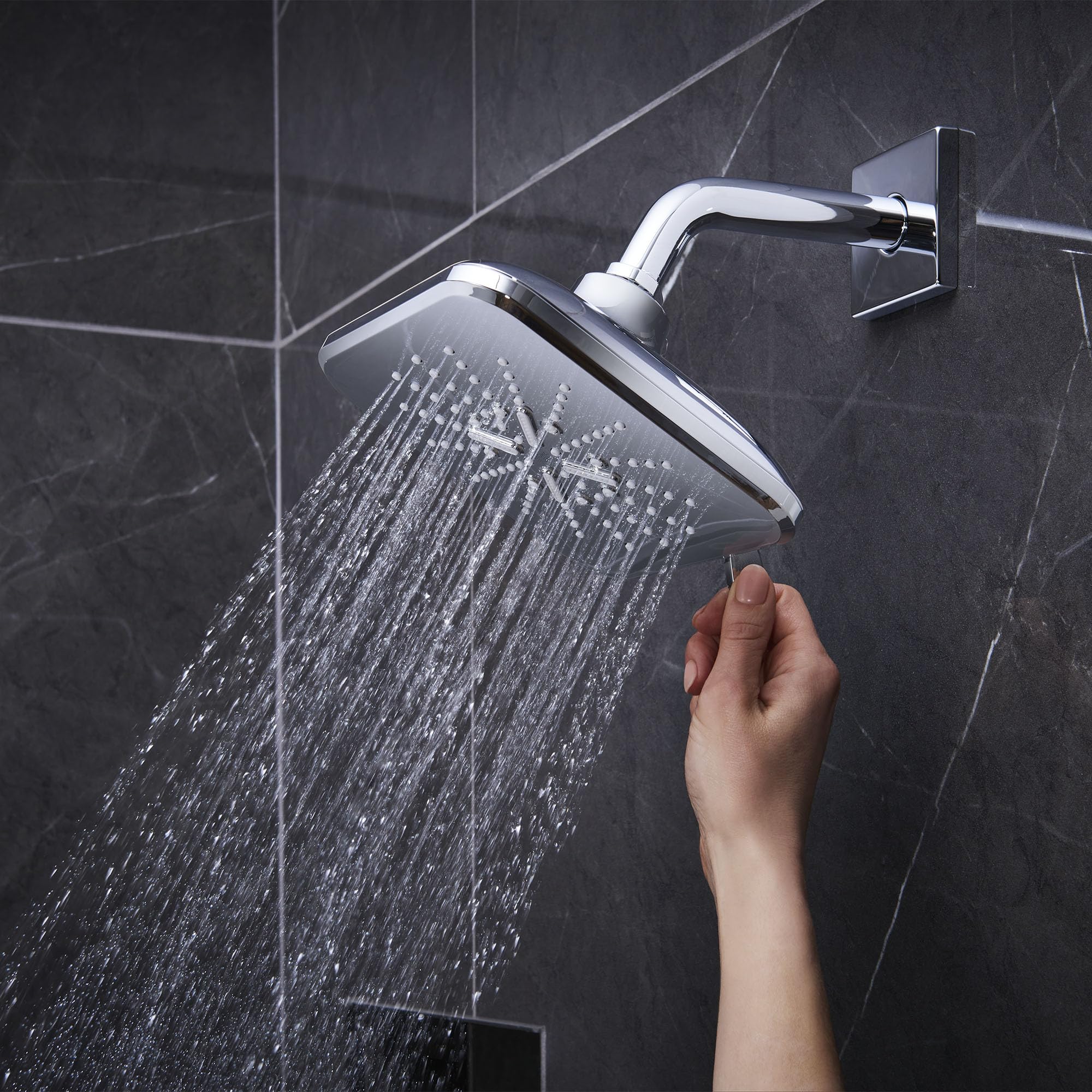 Chrome shower head with curved arm mounted on dark tile wall, dispensing water with hand reaching toward controls below.