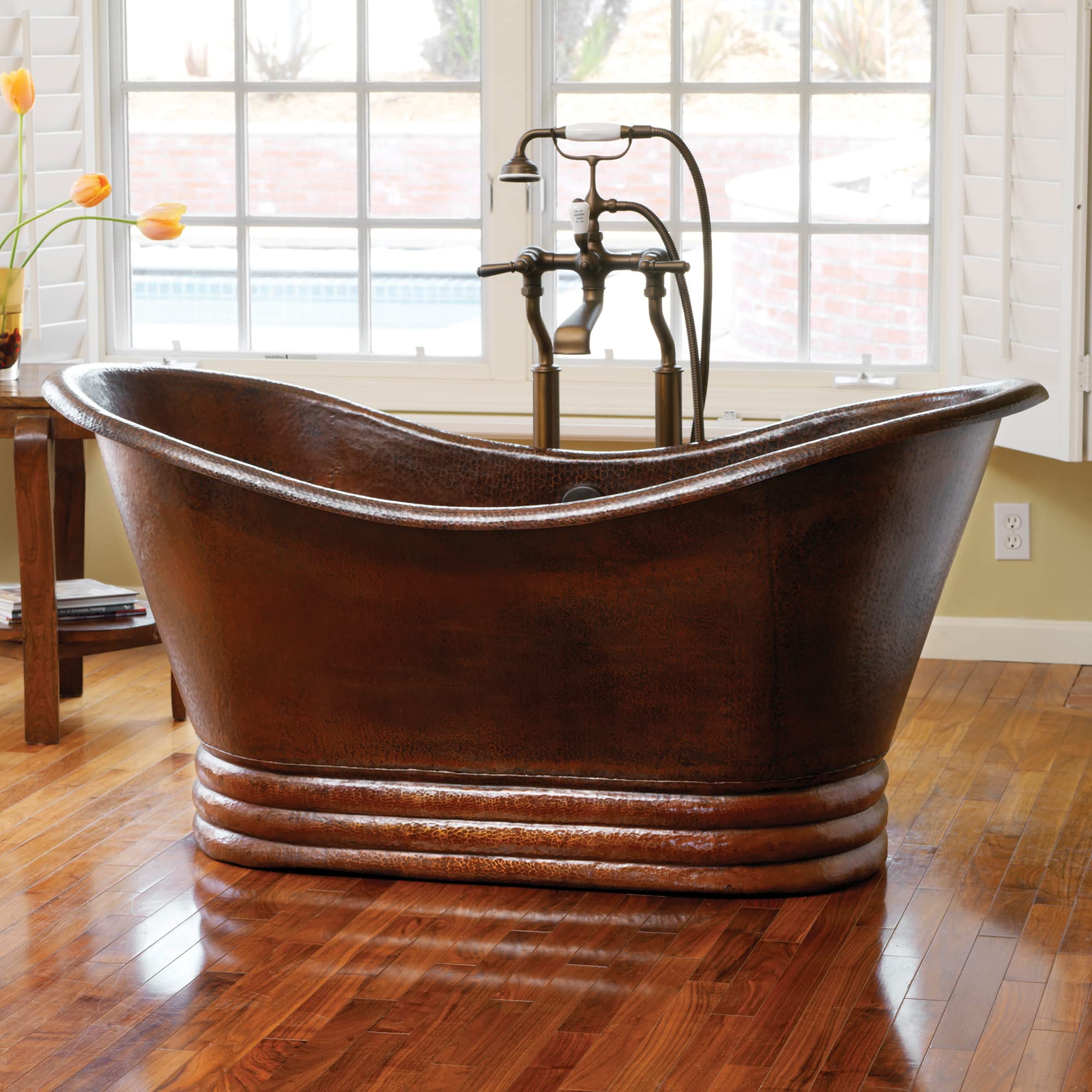 Hammered copper freestanding bathtub with curved rim and decorative base bands, paired with bronze floor-mounted faucet.