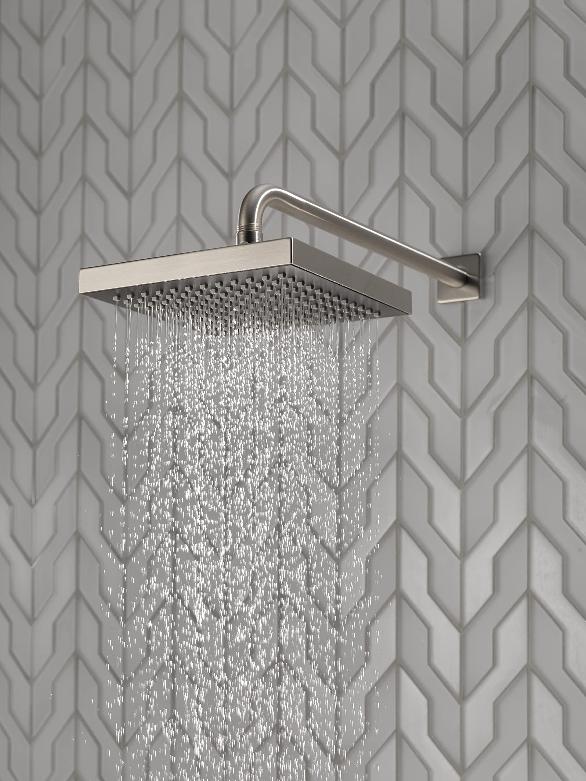 Stainless steel raincan shower head mounted on wall with curved arm, dispensing water against textured tile background.