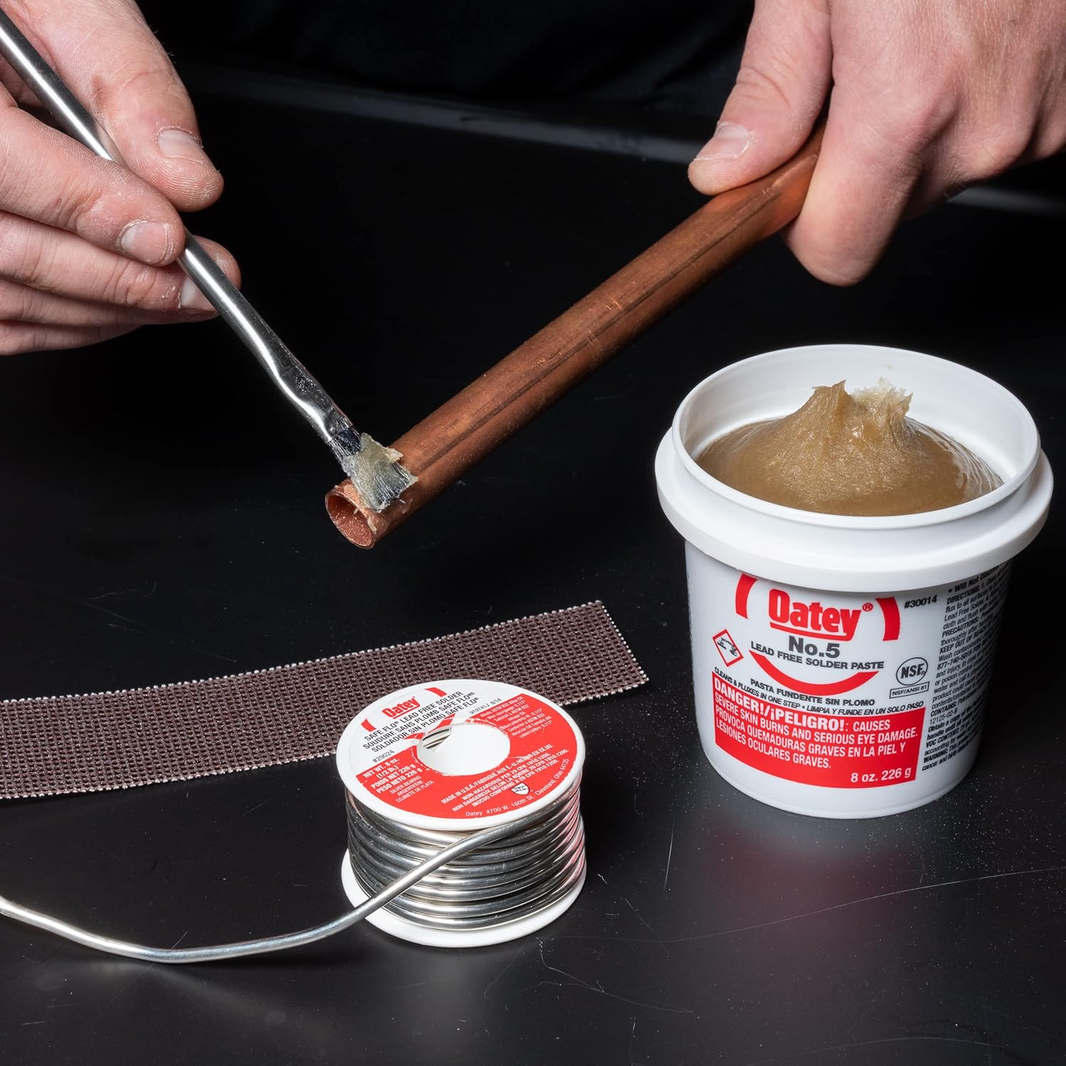 Oatey No. 5 paste flux in white container with red label, displayed with solder spool and copper pipe being soldered.