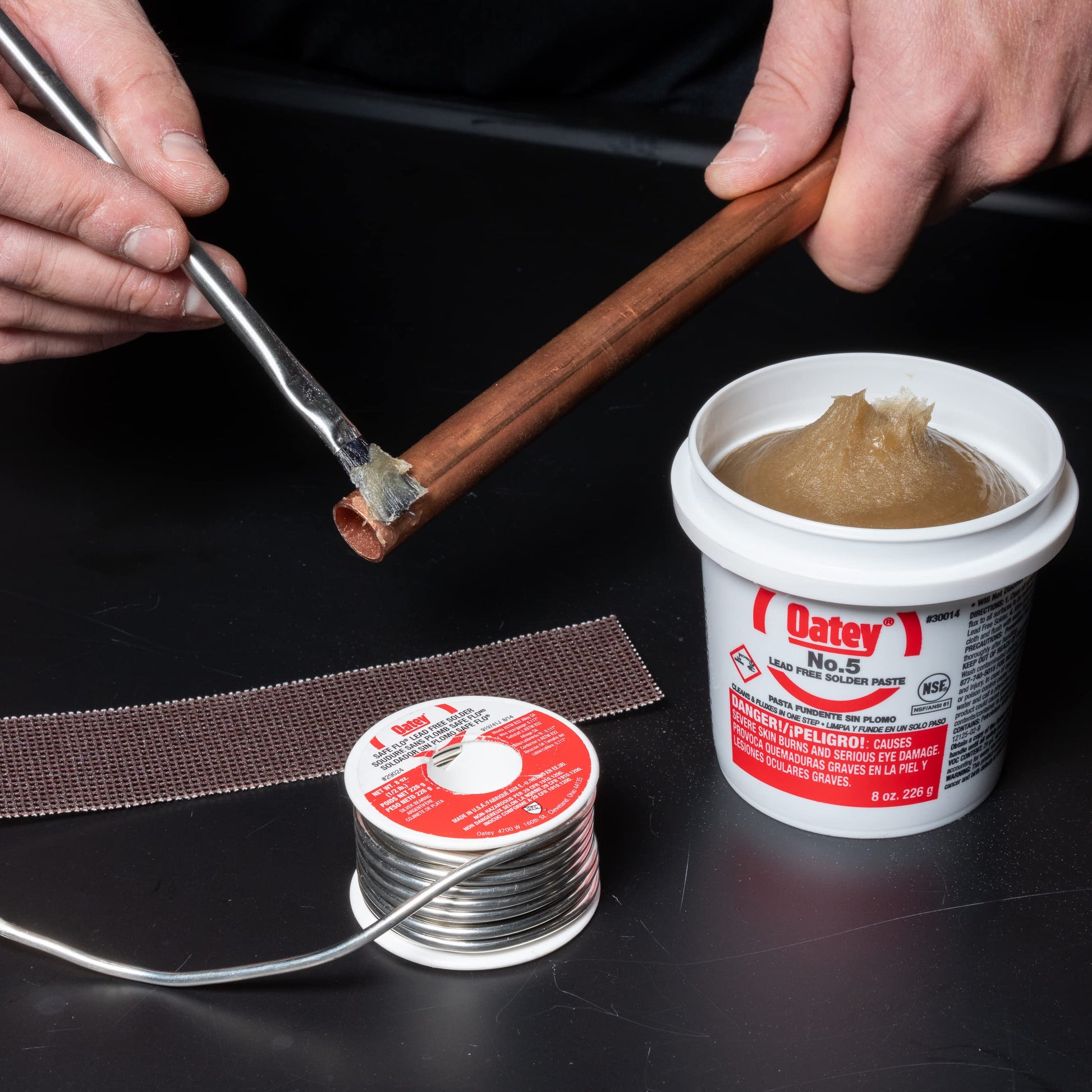 Solder spool with silver-gray wire, flux paste in white container, and copper pipe being soldered with iron tool.
