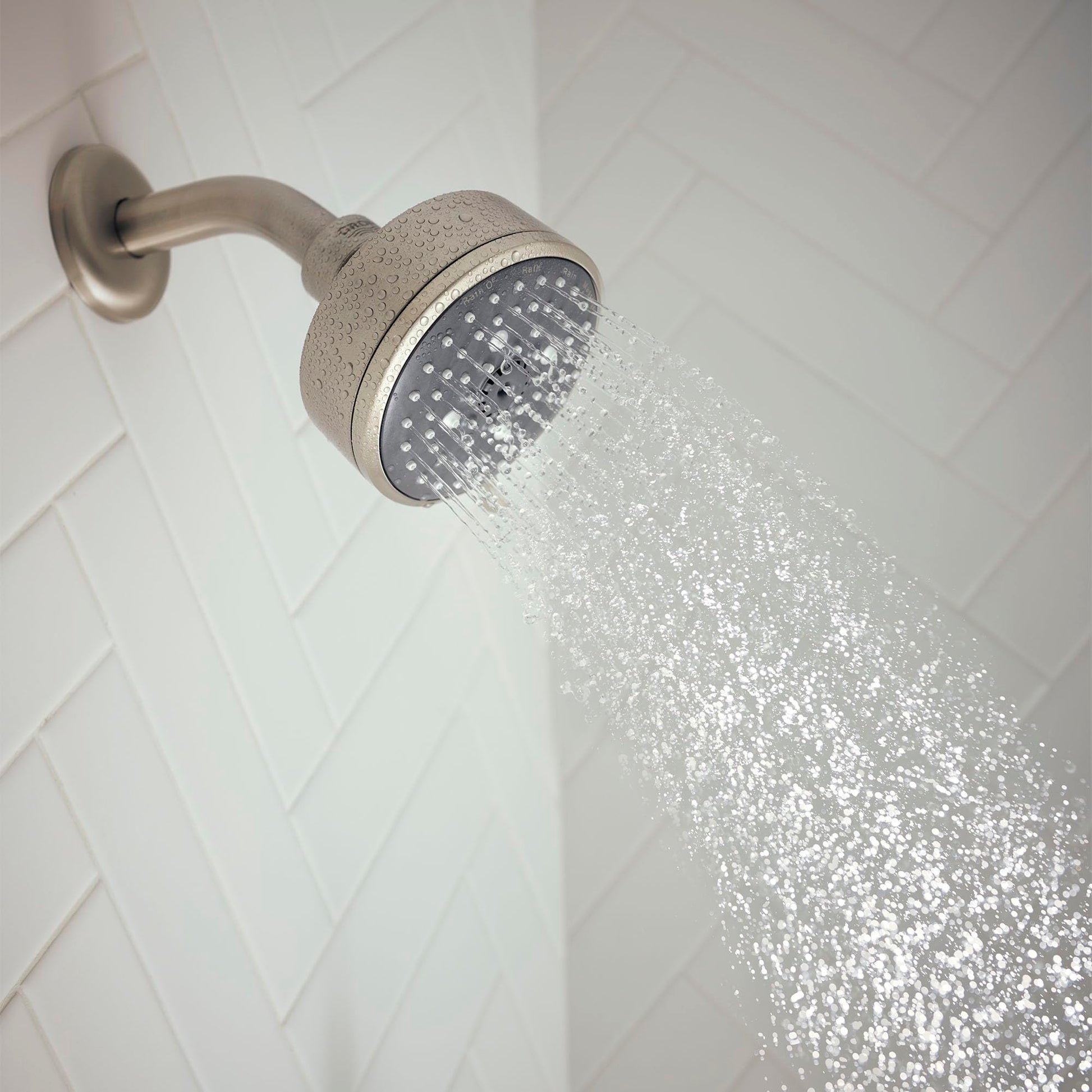 Brushed nickel shower head with perforated face mounted on curved arm, dispensing water spray against white tiled wall.