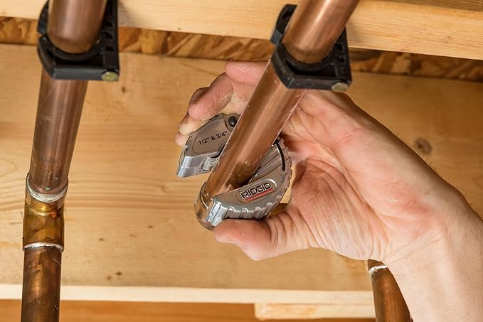 Hands holding RIDGID tubing cutter against two copper pipes mounted horizontally on wooden surface.